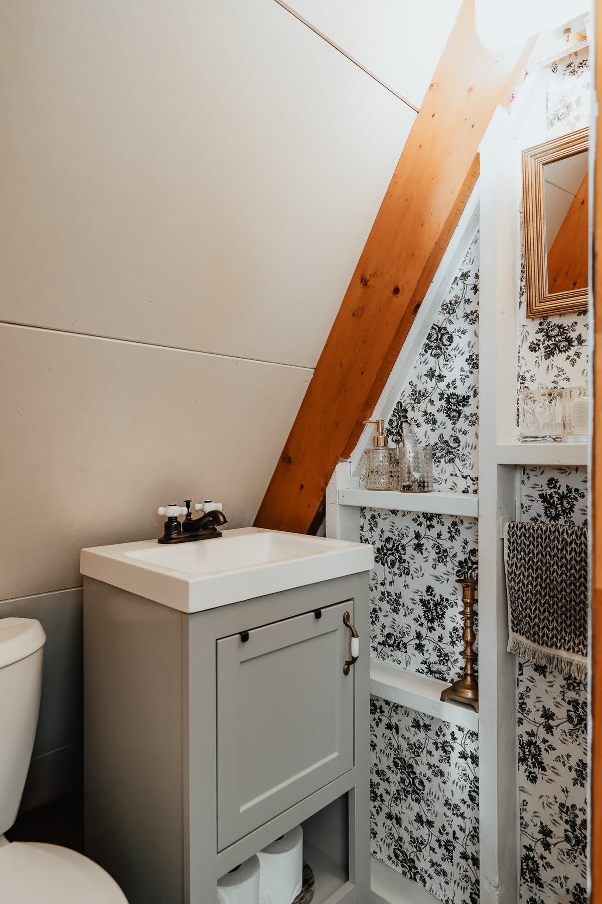 A compact A-frame bathroom features a light gray vanity with a white sink, black and white floral wallpaper, and a small mirror, with exposed wooden beams accentuating the sloped ceiling.