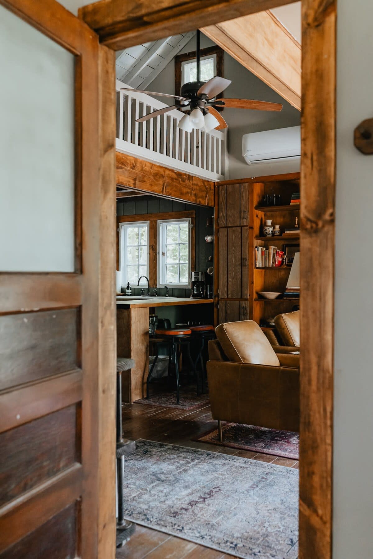 A view through a rustic wooden doorway into the open-concept living area and kitchen, showcasing a leather armchair, a ceiling fan in the loft, and the kitchen's wooden countertops.
