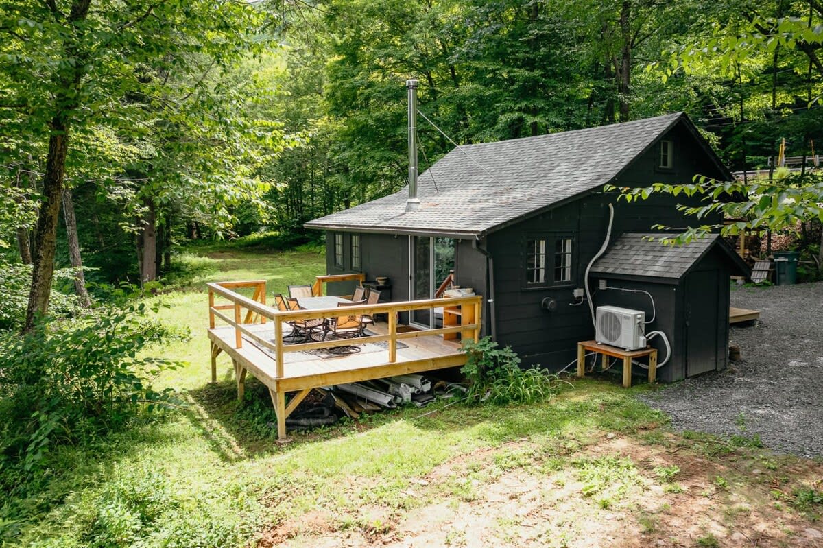 The black cabin from a side view, showcasing a spacious wooden deck with outdoor seating, surrounded by dense green trees and a gravel path leading to the entrance.