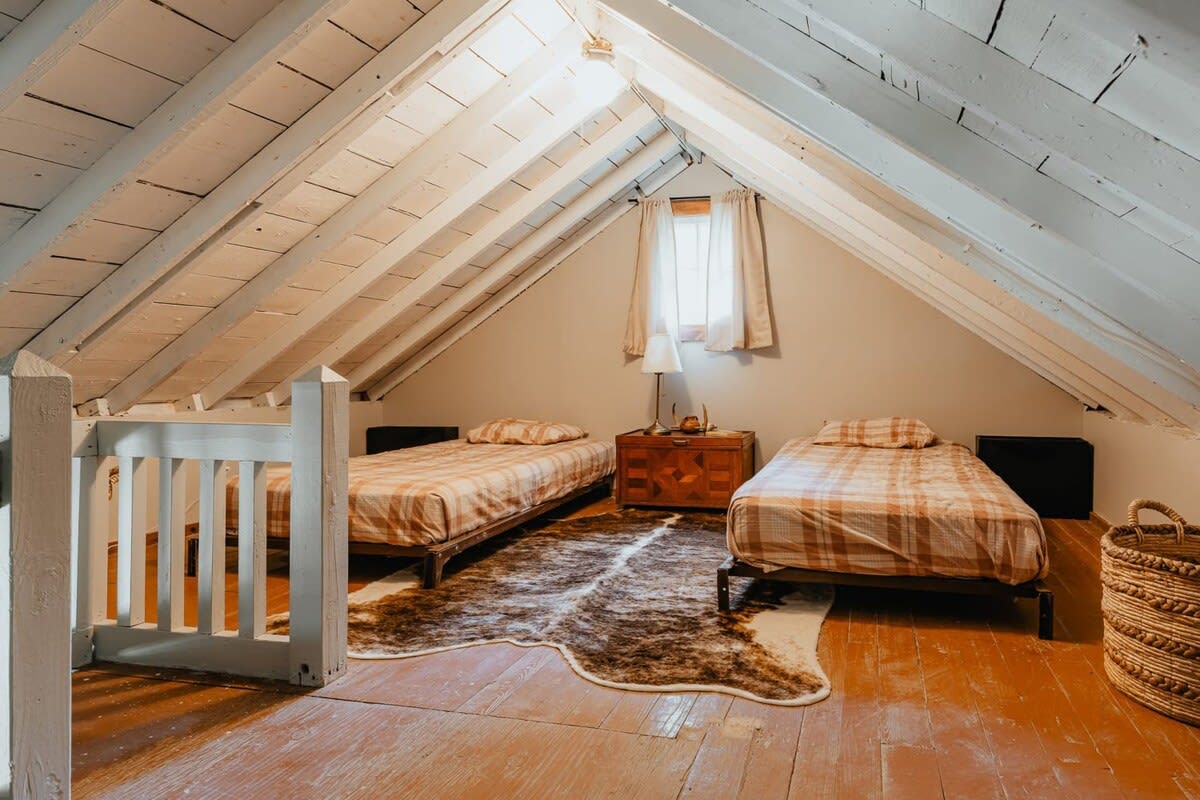 An attic bedroom featuring two twin beds with plaid bedding, set on a light wooden floor with a cowhide rug. The sloped ceiling has exposed white-painted beams, and a small window provides natural light.