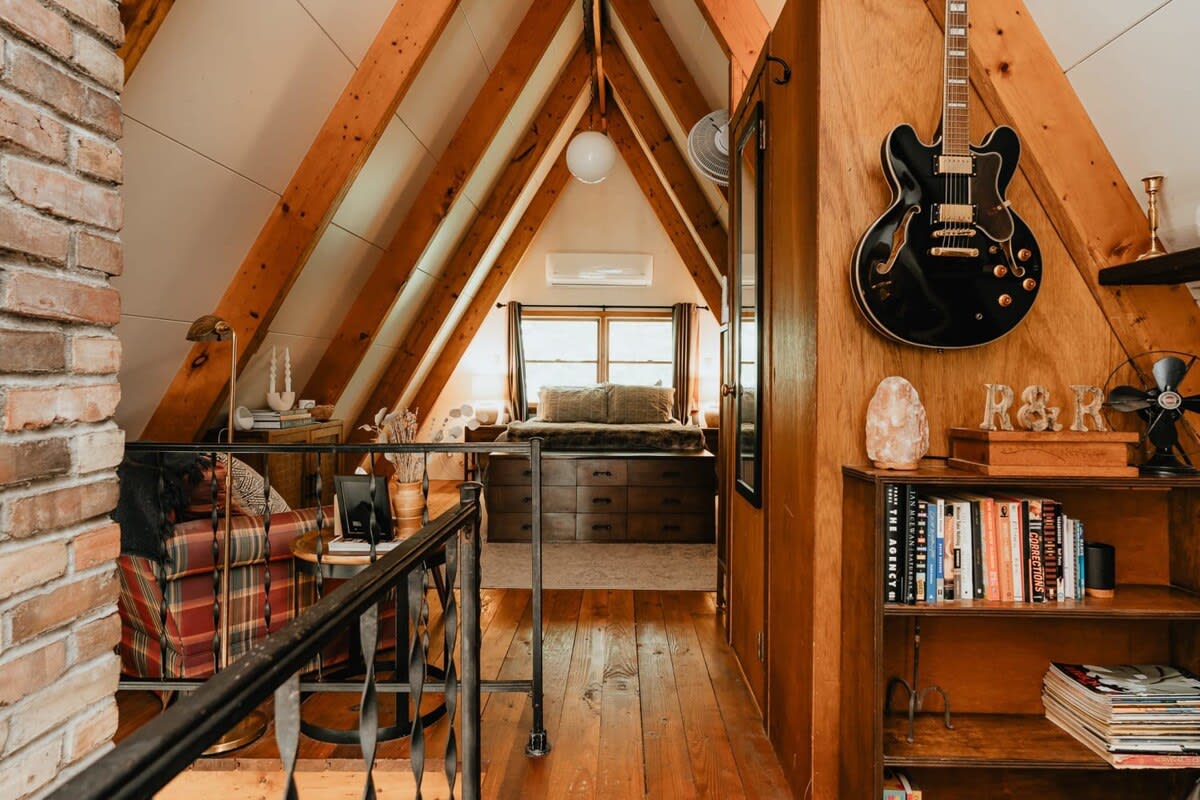 View from a loft showing a bedroom area with a large bed, a wooden dresser, and an air conditioning unit, framed by exposed A-frame beams. A black electric guitar hangs on a wooden wall next to a bookshelf.