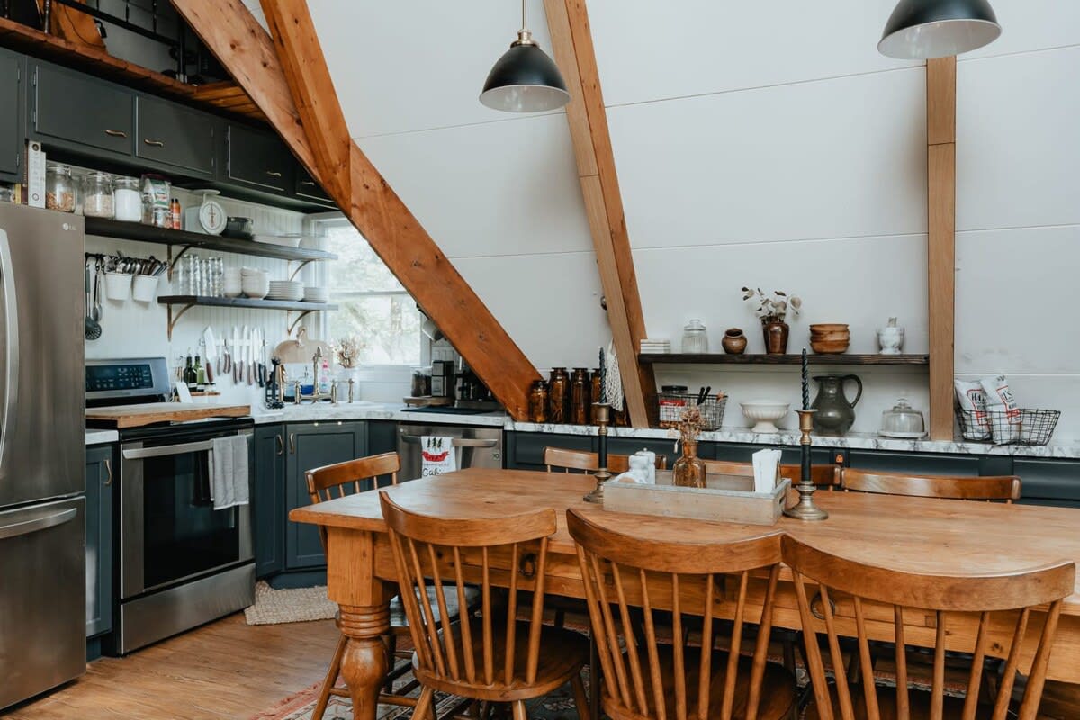 Rustic dining area with a large wooden table and spindle-back chairs, featuring exposed wooden beams and a view into a dark-cabinet kitchen with open shelving.