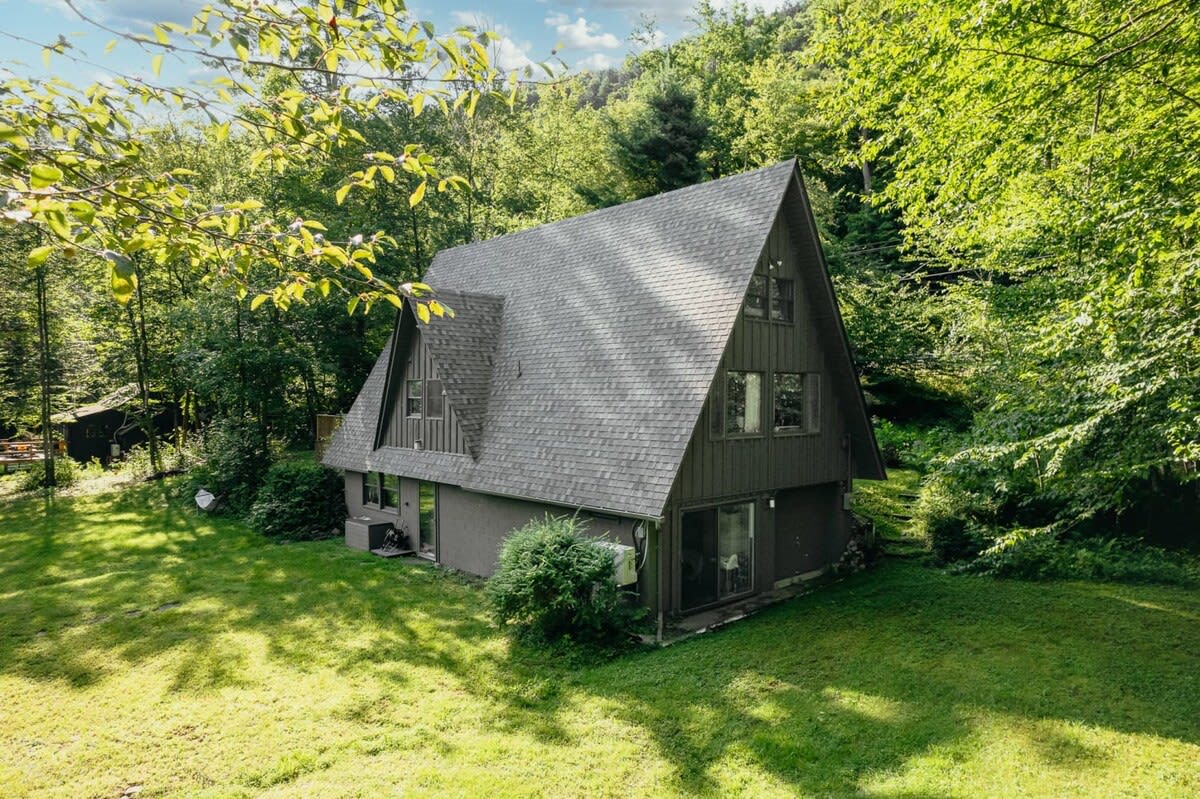 An aerial view of a dark-colored A-frame cabin with a gray shingled roof, nestled amidst a vibrant green forest, showcasing its unique architectural style and secluded setting.