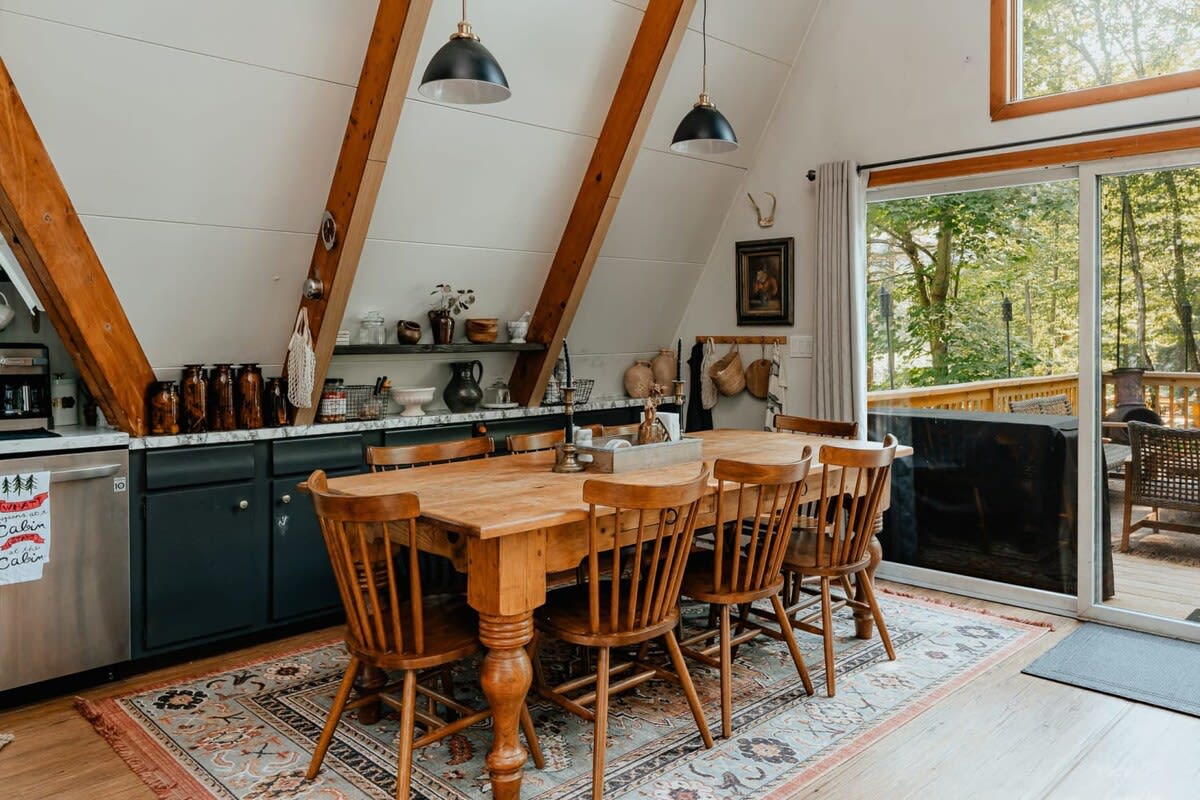 A rustic wooden dining table with six spindle-back chairs sits on a patterned rug, beneath two black pendant lights, with a kitchen counter and exposed wooden beams in the background.