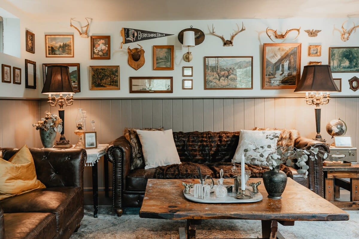 A rustic living room with dark leather sofas, a wooden coffee table, and a wall adorned with numerous framed pictures, antlers, and sconces, creating a cozy, lodge-like atmosphere.
