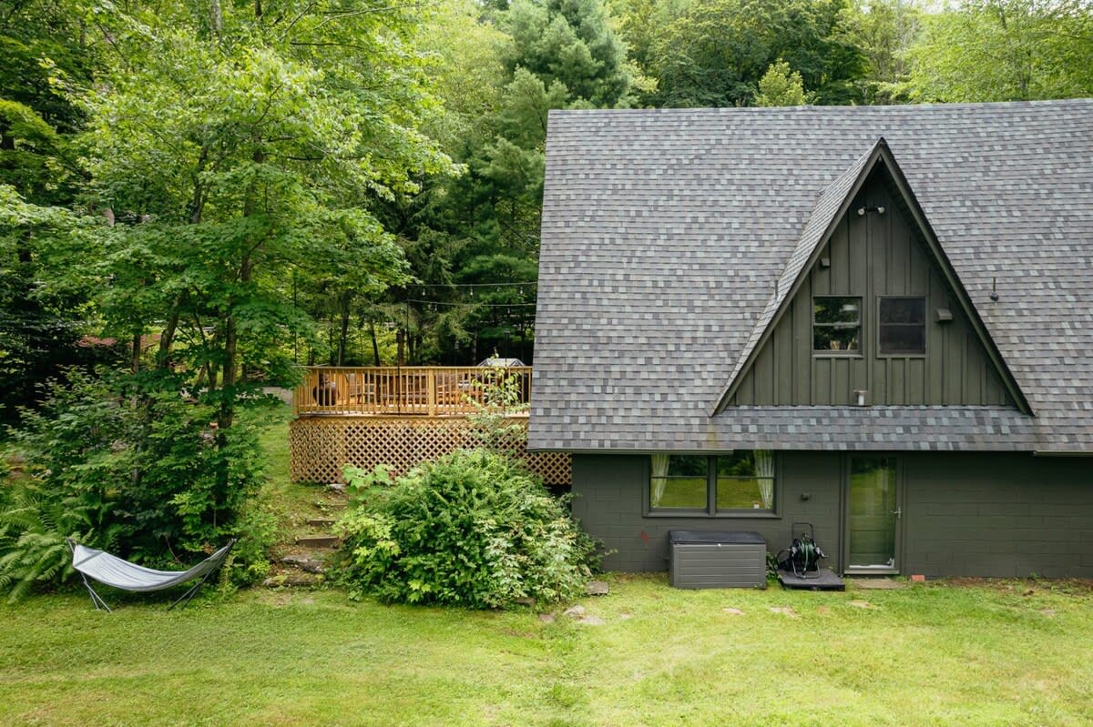 The back exterior of the A-frame cabin, showing a side view of the shingled roof and dark siding, with a wooden deck visible on the left and a hammock set up in the grassy yard.