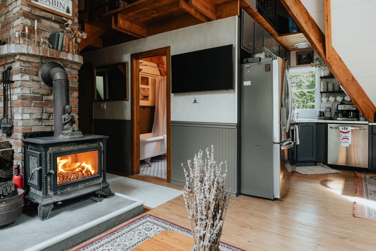 A rustic living space centered around a brick fireplace with a black wood-burning stove, offering a glimpse into a bathroom with a clawfoot tub and a partial view of the kitchen area.