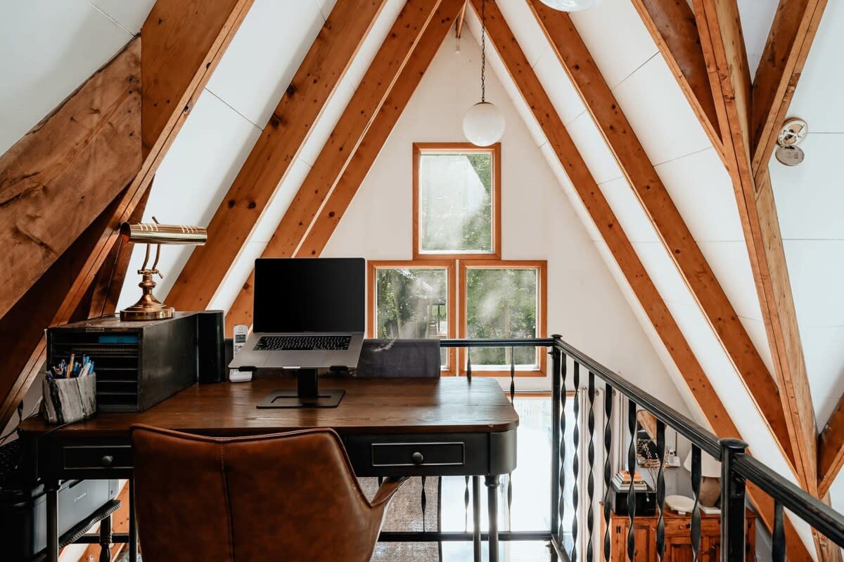 A rustic loft office space with a dark wood desk, leather chair, and laptop, set against a backdrop of multiple windows and exposed A-frame beams. A black metal railing overlooks the lower level.