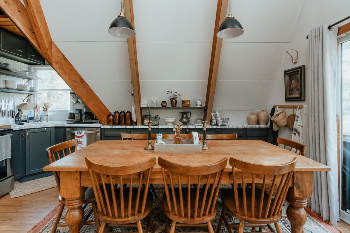 A wide shot of the dining area highlights the large wooden table and chairs on a patterned rug, with dark kitchen cabinets and open shelving visible on both sides, and exposed wooden beams overhead.
