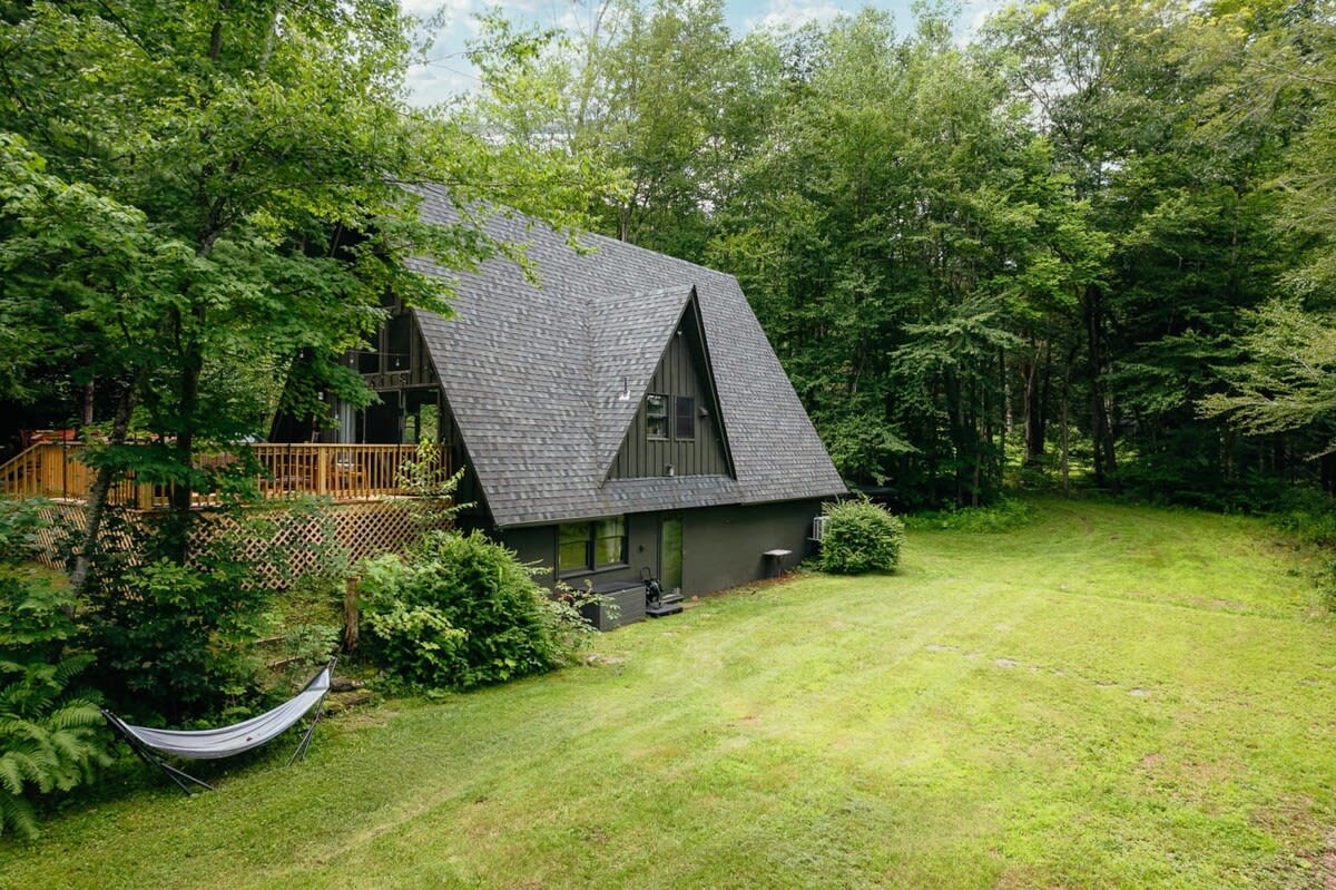 A dark green A-frame cabin with a gray shingle roof is nestled among lush green trees, featuring a wooden deck on the left side and a white hammock in the grassy front yard.