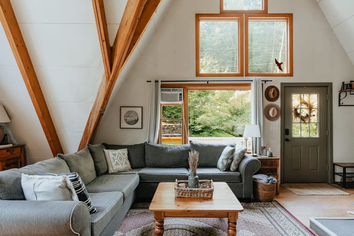 A large gray sectional sofa with throw pillows sits on a patterned rug, facing a rustic wooden coffee table, with exposed wooden beams and large windows offering views of the surrounding trees.