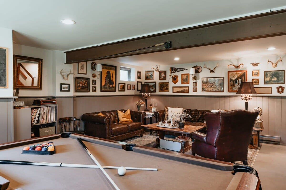 A cozy game room featuring a pool table in the foreground and a comfortable seating area with leather sofas and an armchair in the background, adorned with a gallery wall of framed pictures and antlers.
