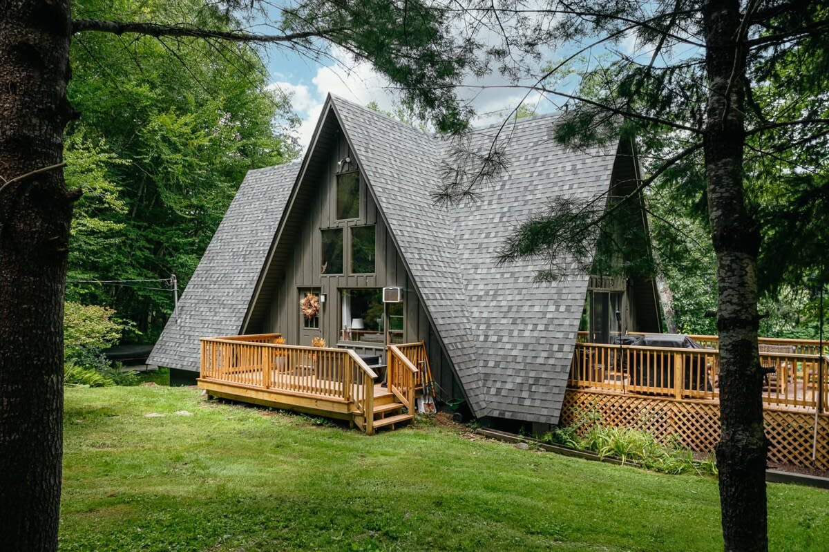 The front exterior of a dark A-frame cabin with a prominent wooden deck and railing, surrounded by lush green trees and a well-maintained lawn, highlighting its rustic charm.