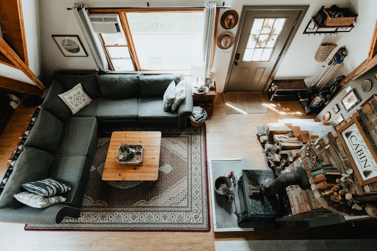 An aerial view of a cozy living area with a grey sectional sofa, wooden coffee table, and a brick fireplace with a wood-burning stove. Light hardwood floors and an area rug define the space.