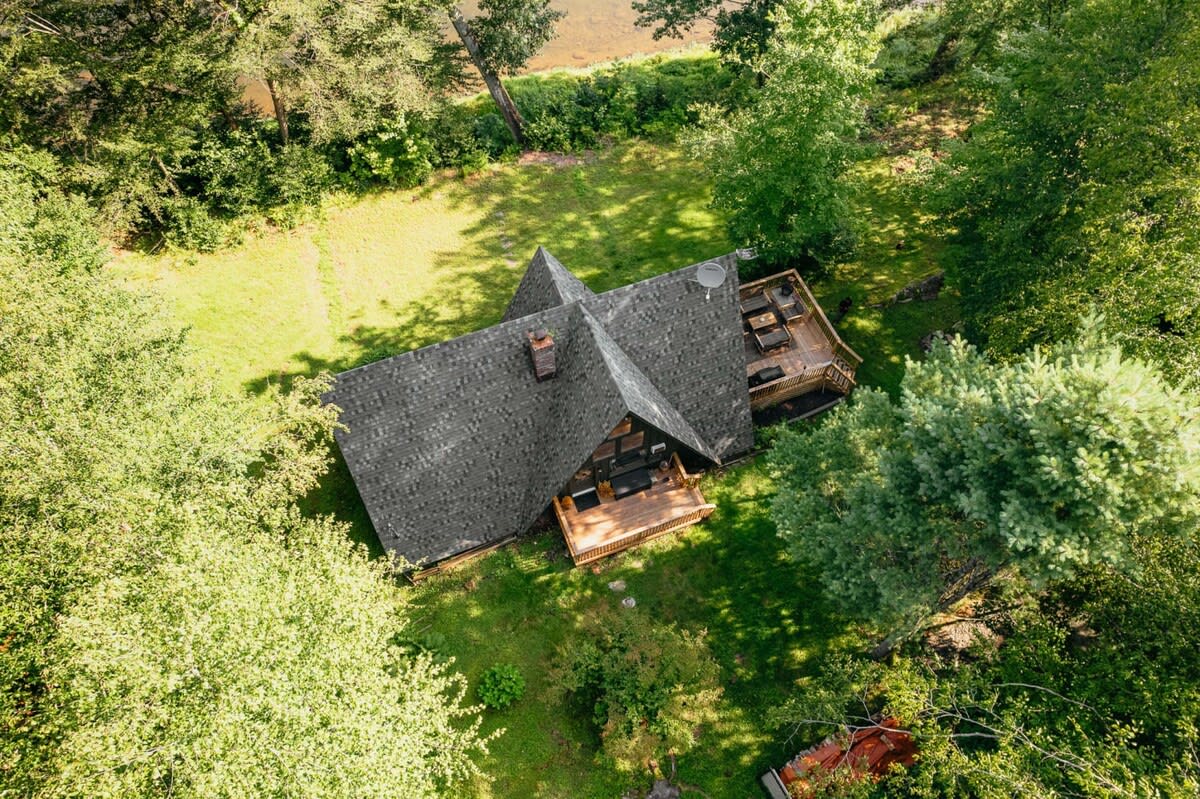 An aerial view shows the dark gray A-frame cabin with its shingle roof, surrounded by dense green forest, with a glimpse of a river or body of water in the upper right background.