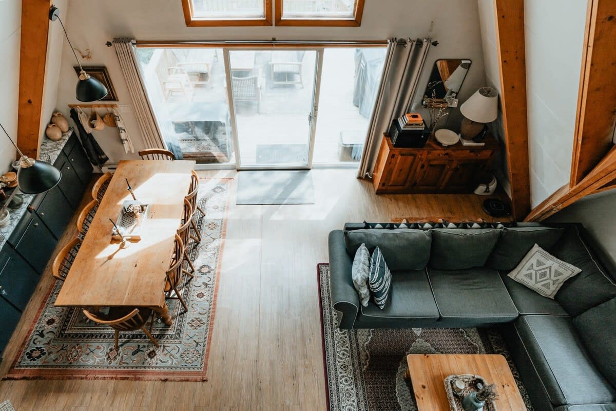 An aerial view of an open-concept living and dining area with light hardwood floors, featuring a large grey sectional sofa, a wooden coffee table, and a long wooden dining table with chairs. Large glass doors lead to an outdoor area.
