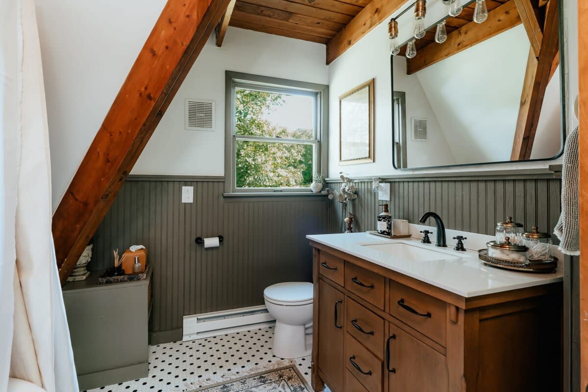 A rustic bathroom featuring a dark wood vanity with a white countertop, black fixtures, and beadboard wainscoting. The sloped ceiling with exposed wooden beams adds to the cabin aesthetic.