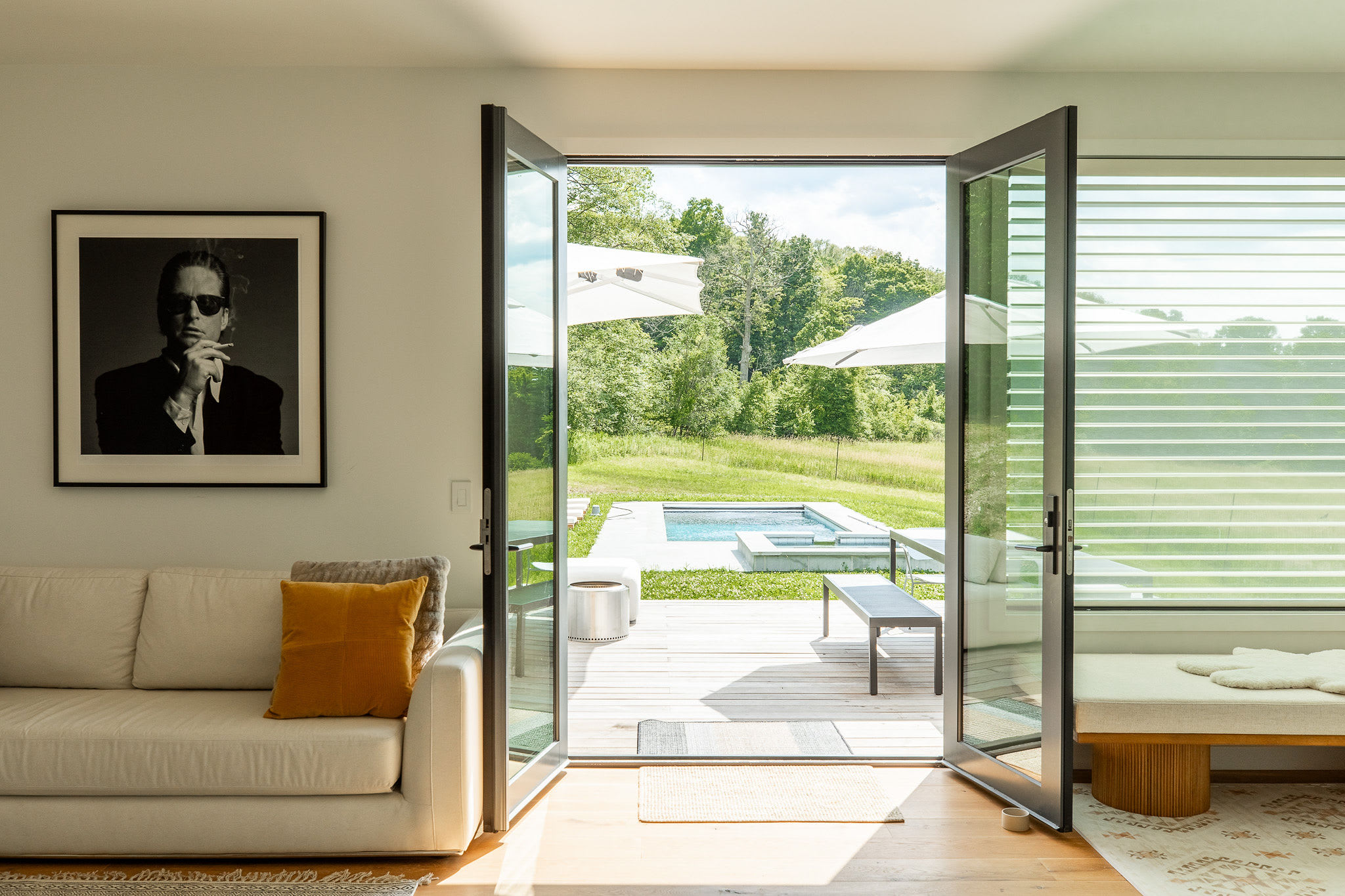 Cozy living area with a light-colored sofa and a framed black and white portrait, featuring double glass doors that open to a wooden deck with a swimming pool and lush green landscape views.