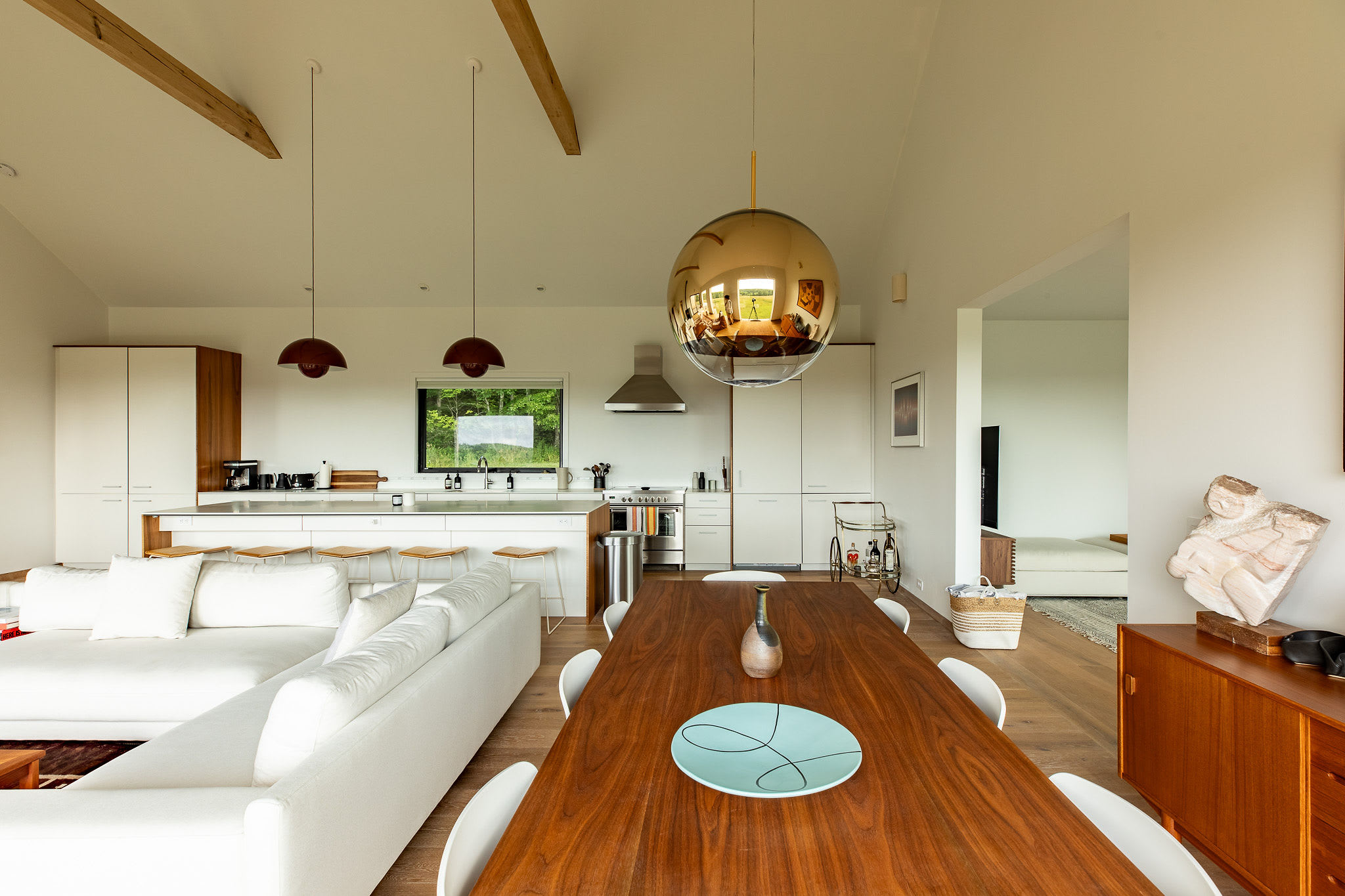An open-concept dining area features a long wooden dining table with white chairs, illuminated by a large spherical gold pendant light. The space connects to a modern kitchen with white cabinets and a living area, all under a vaulted ceiling with exposed beams.