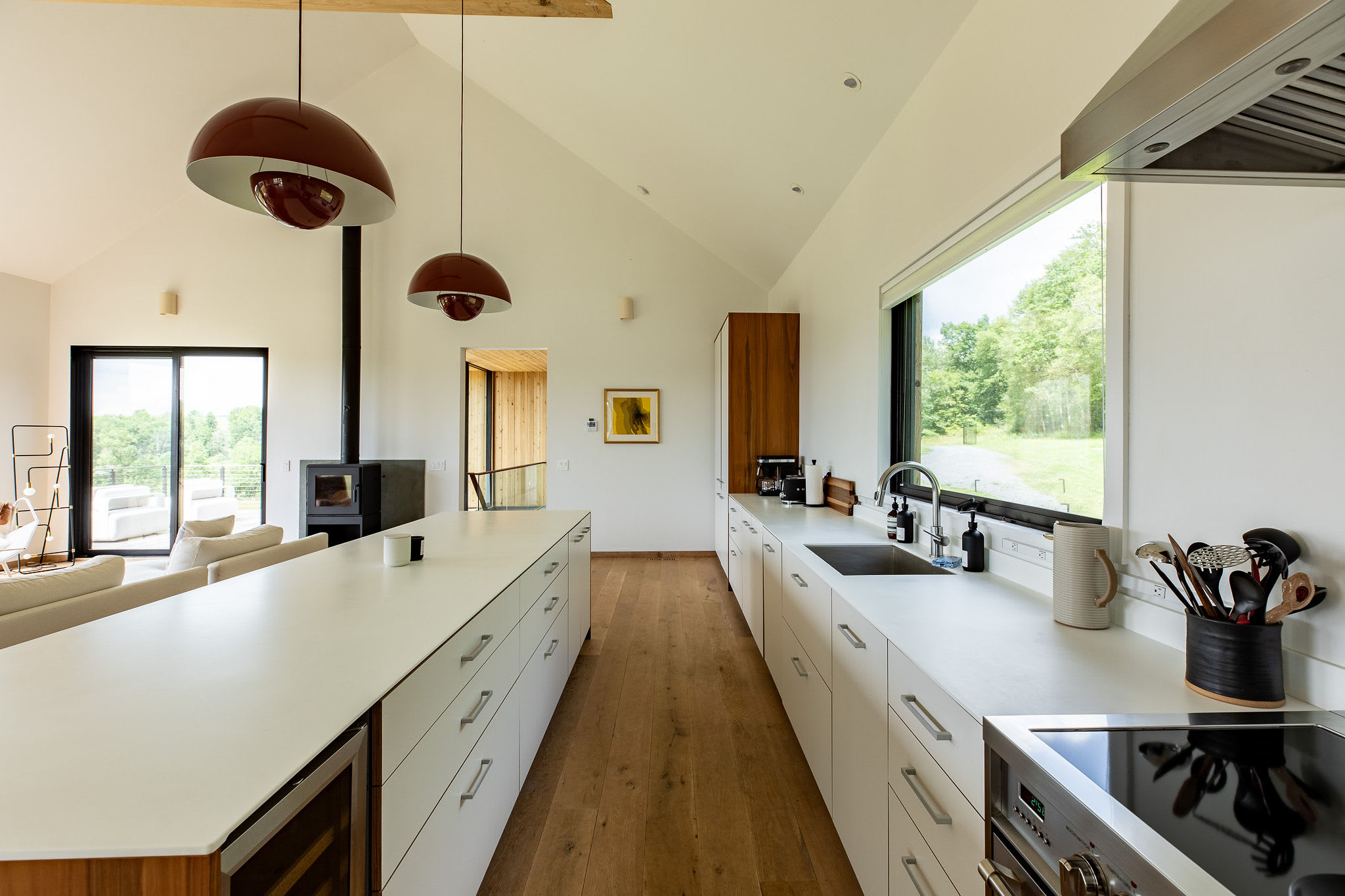 A sleek, open-concept kitchen features a long white island and white perimeter cabinets with wooden accents, facing a modern wood-burning stove and a living area. The space has vaulted ceilings with exposed beams and a large window overlooking green trees.