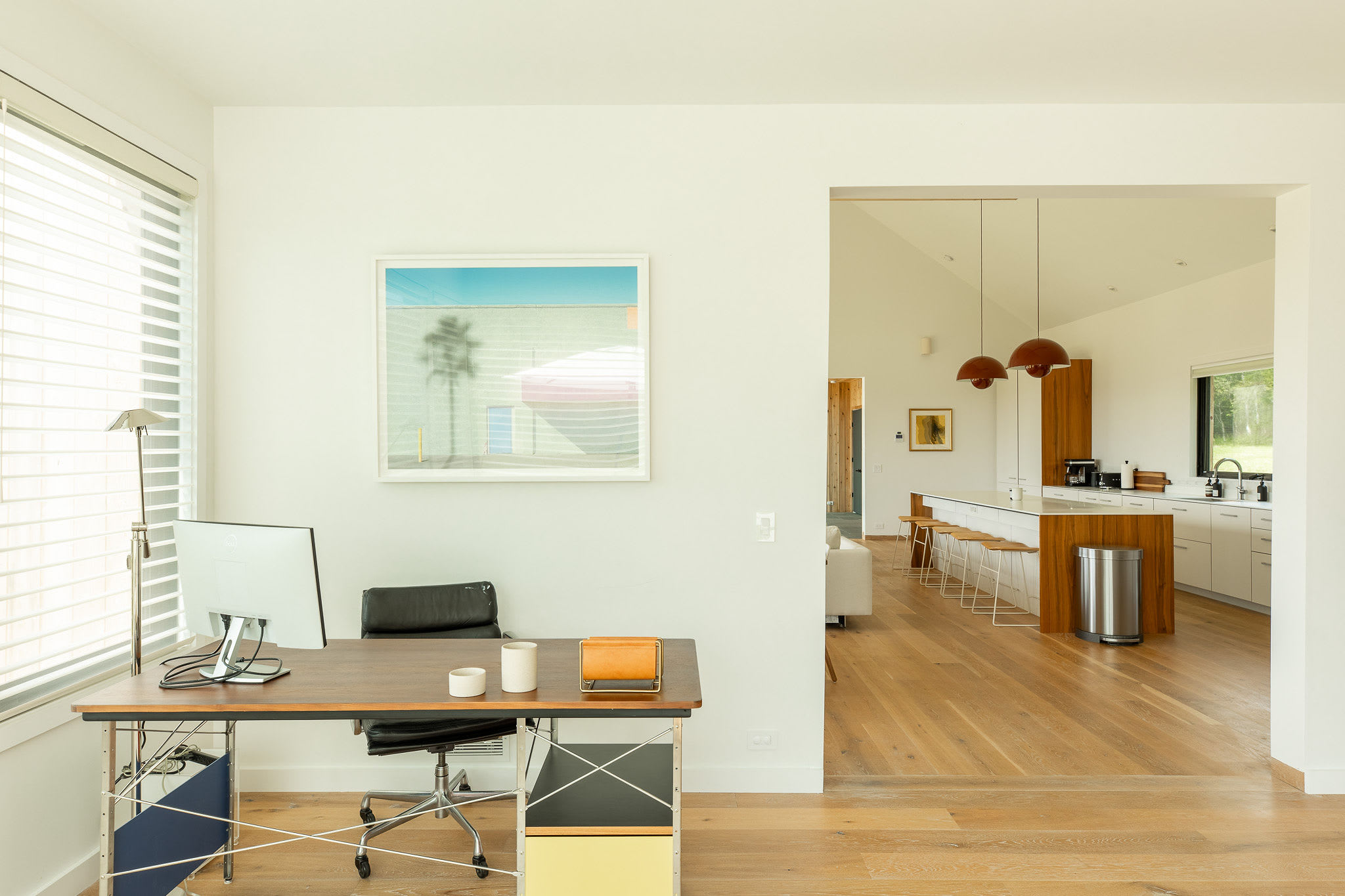 Modern home office with a wooden desk, black office chair, and a large monitor, featuring a framed abstract artwork on the white wall and an open view into a bright kitchen with a large island.