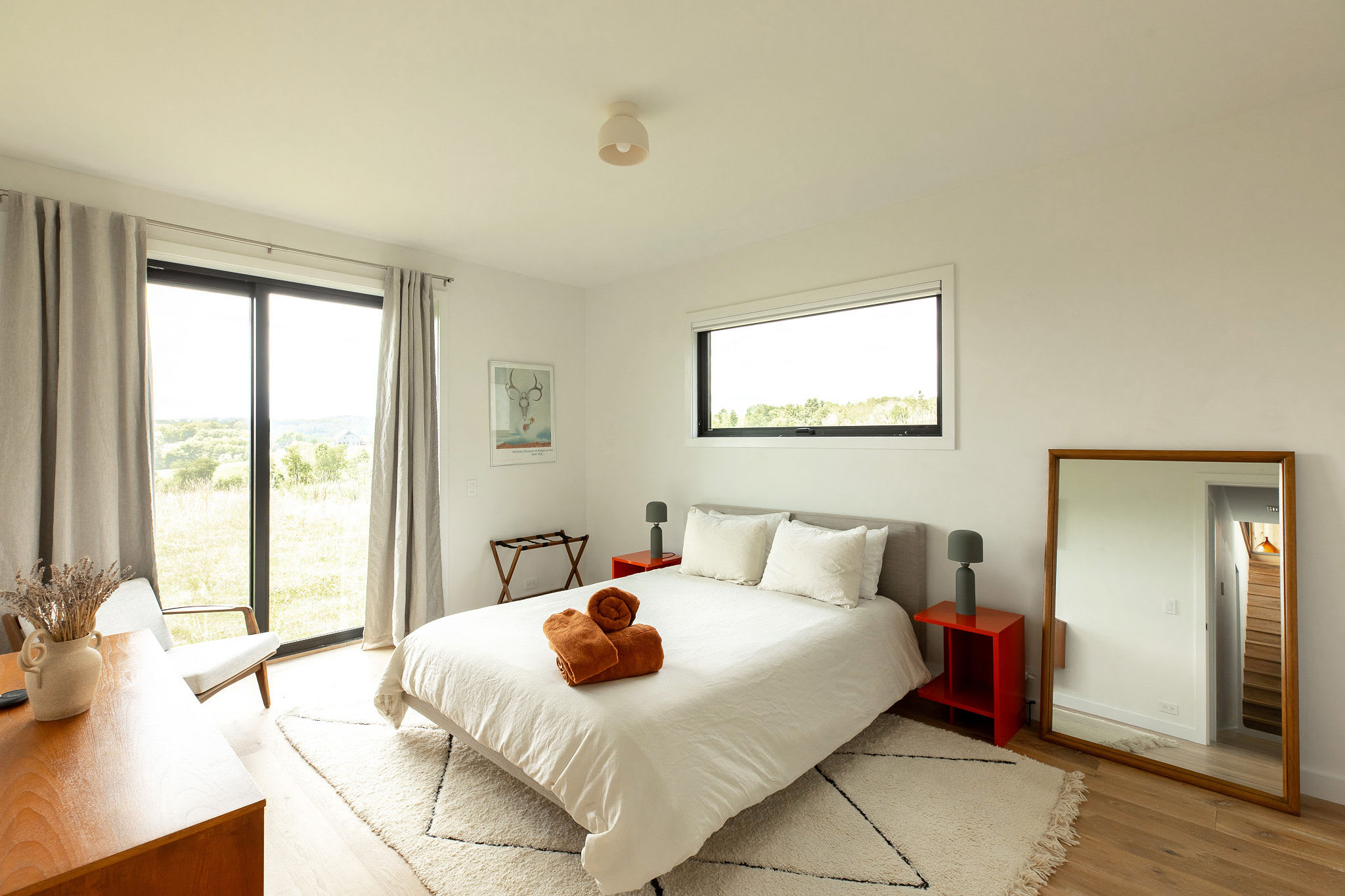 Bright bedroom with a queen bed dressed in white linen and orange towels, flanked by red nightstands with grey lamps. A large sliding glass door and a horizontal window offer expansive views of the green landscape, complemented by a full-length mirror.