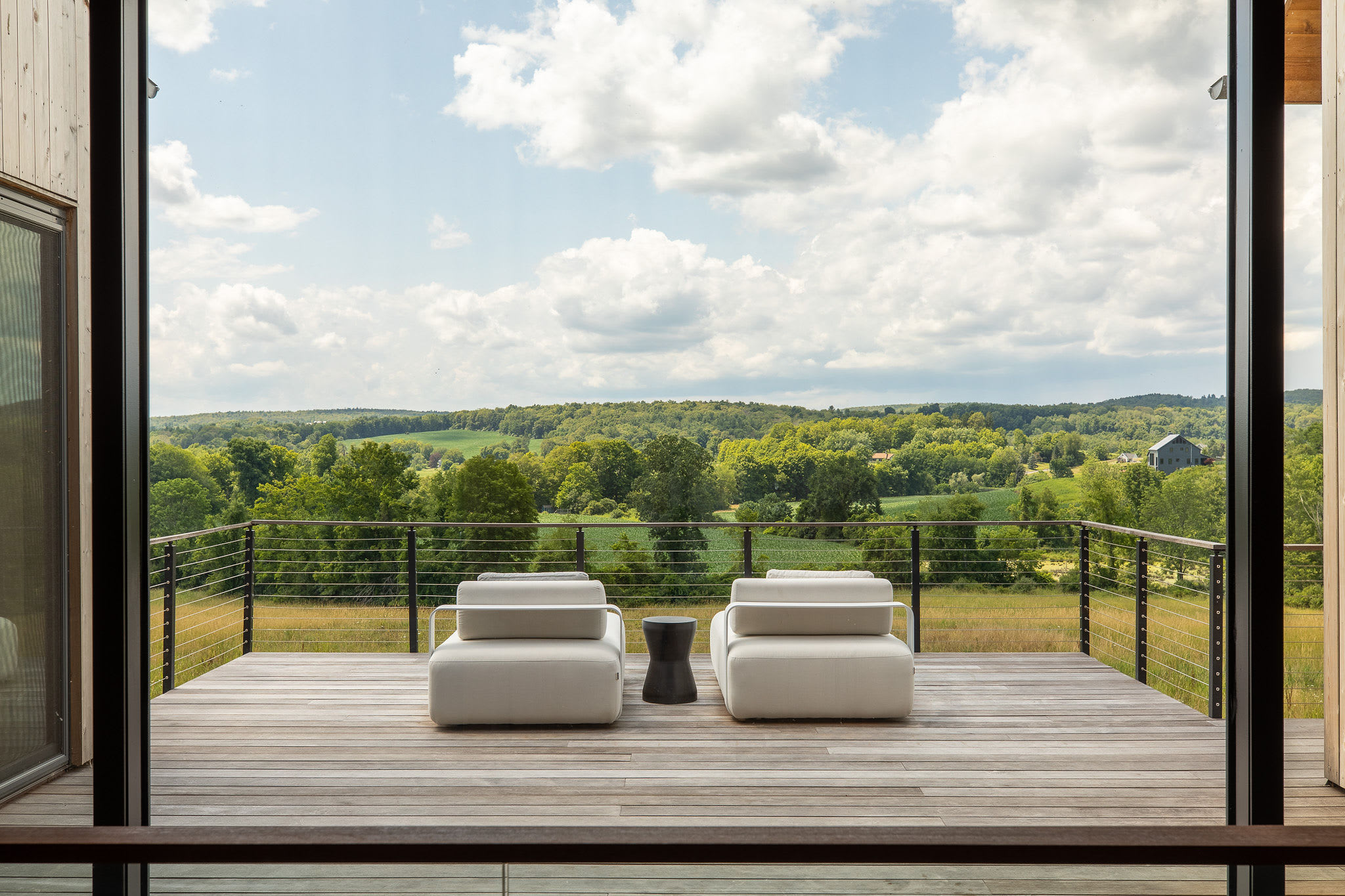 A spacious wooden deck with modern cable railings features two comfortable white outdoor armchairs and a small black side table, offering expansive views of a rolling green landscape under a cloudy sky.