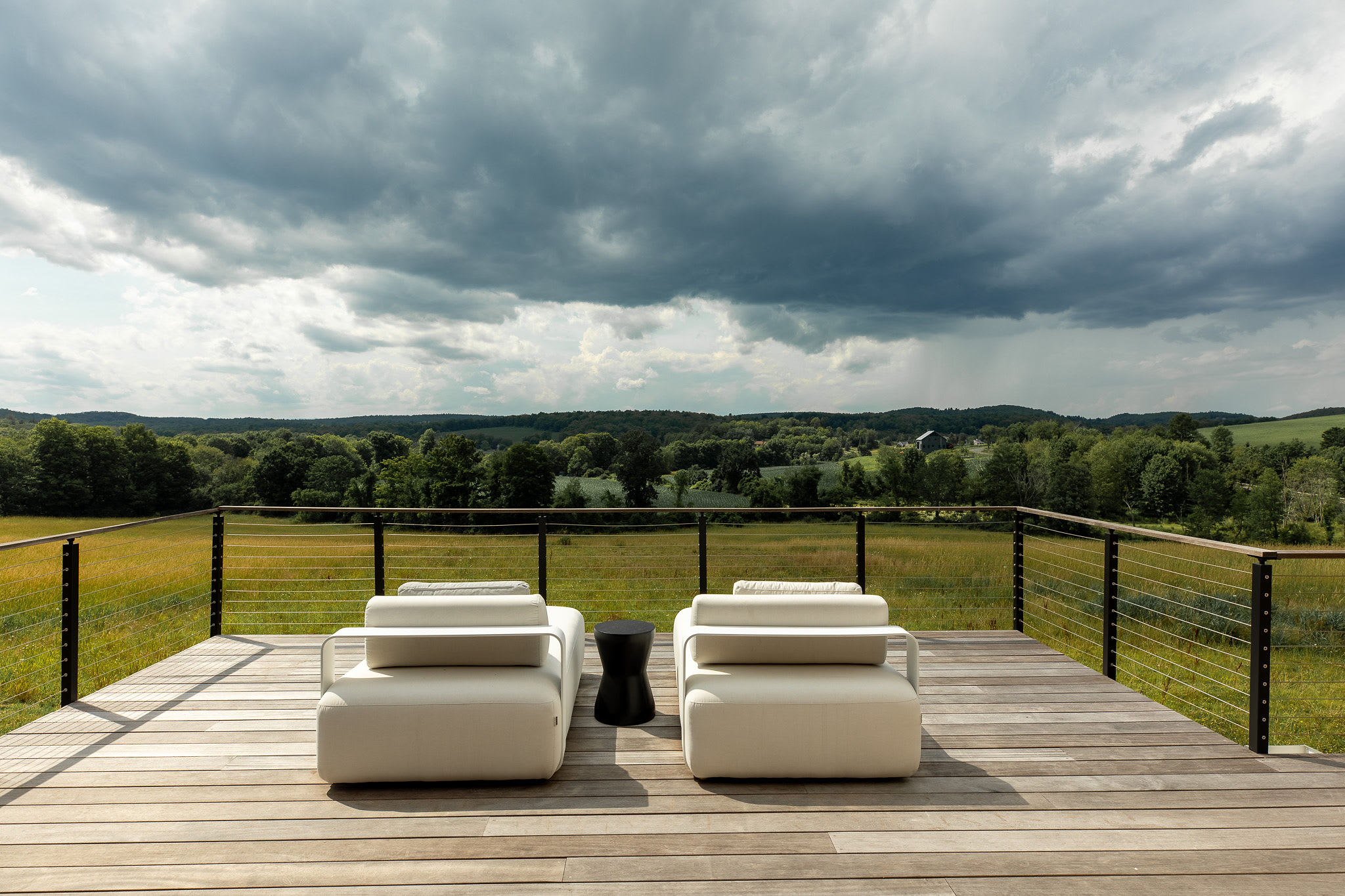 Two modern white outdoor lounge chairs with a small black side table sit on a light wooden deck, offering panoramic views of rolling green hills and distant trees under a dramatic, cloudy sky.