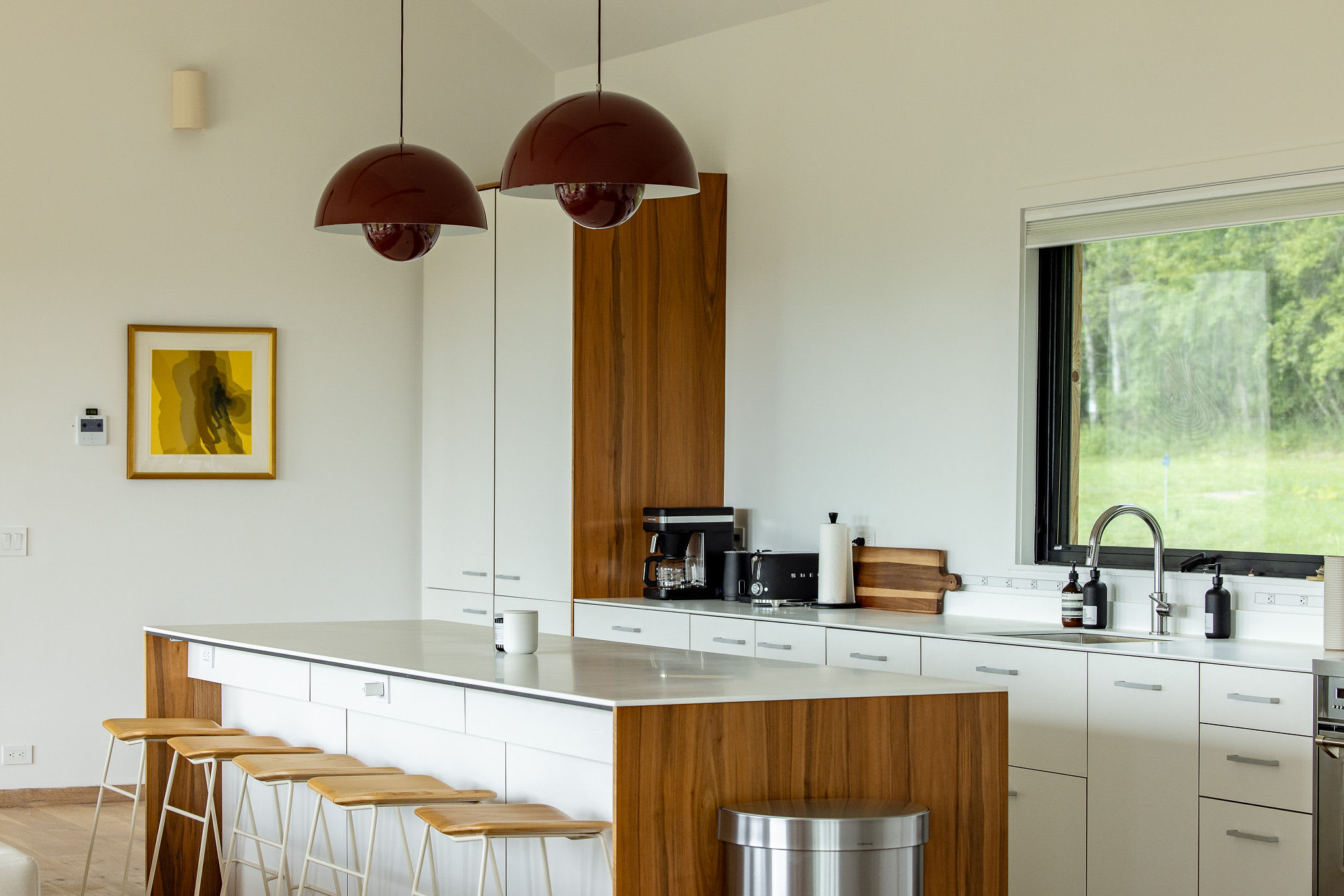 A modern kitchen features a long white island with wooden accents and four bar stools, complemented by white cabinetry and stainless steel appliances. Two large red pendant lights hang above the island, and a window offers a view of green foliage.