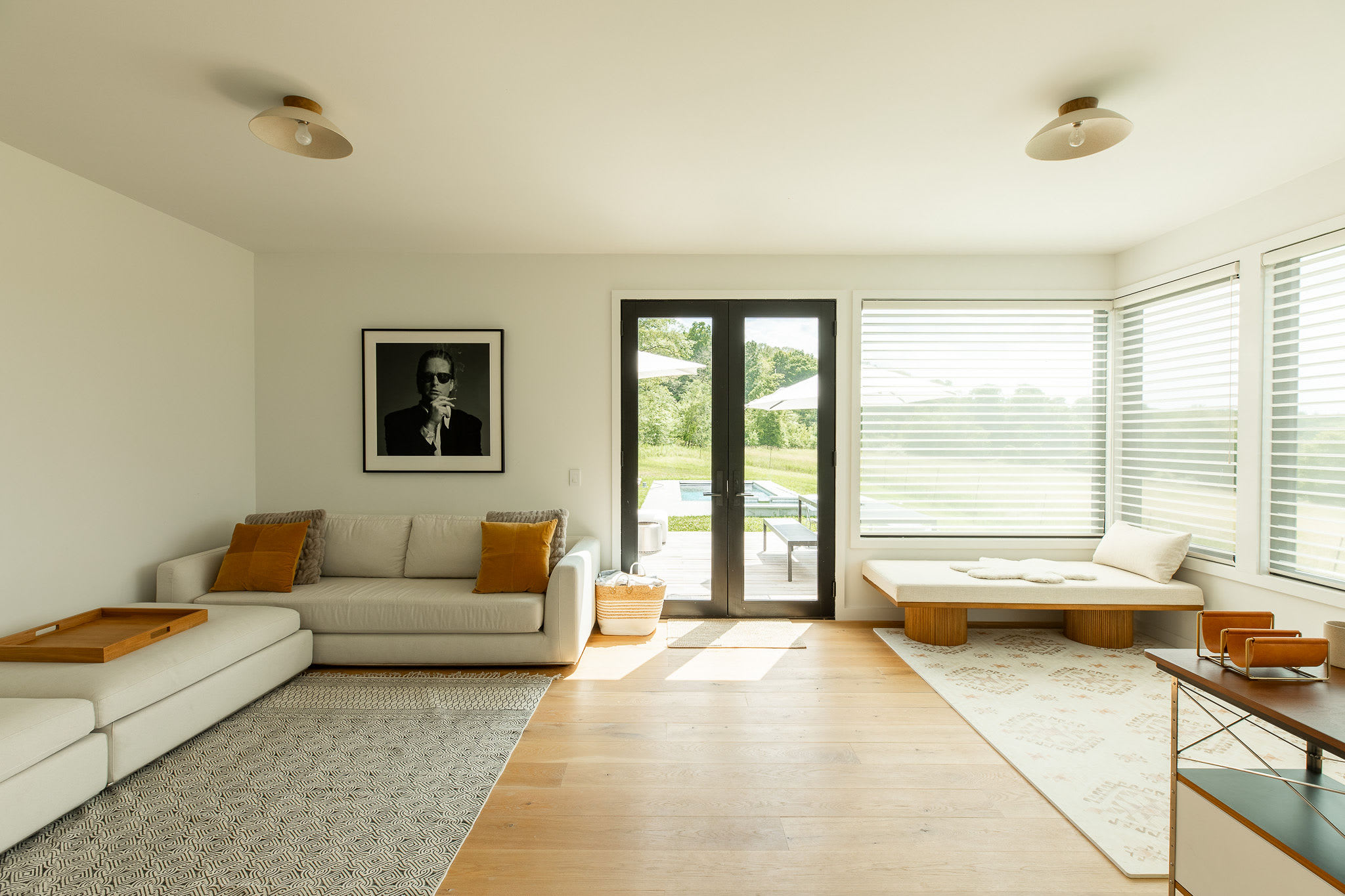 A bright living room features a large L-shaped sectional sofa with mustard yellow pillows, light wood floors, and a large black-framed artwork. Floor-to-ceiling glass doors open to an outdoor deck with a pool, and large windows with blinds offer views of the surrounding landscape.