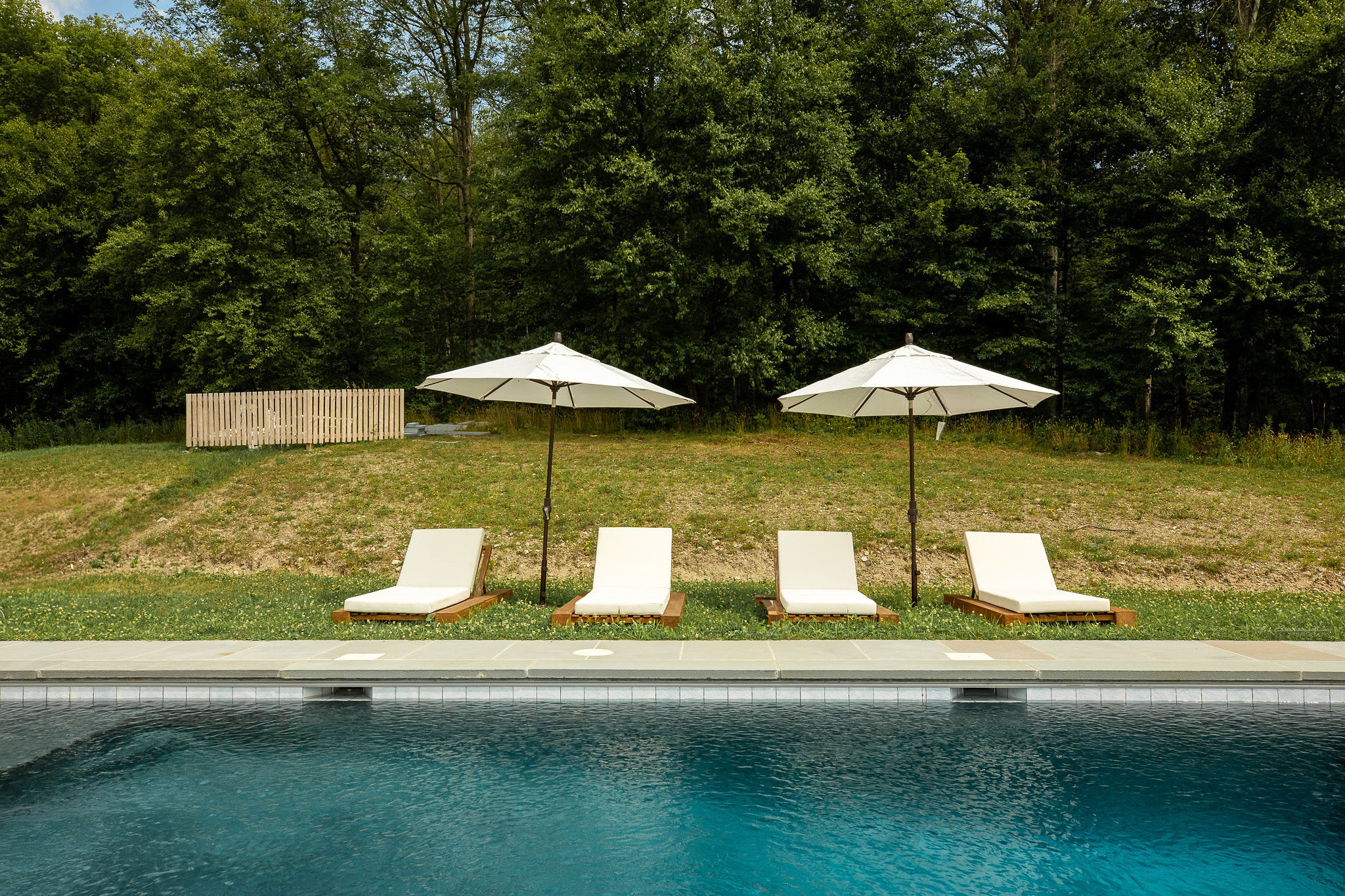 Four white lounge chairs with wooden bases are arranged beside a rectangular inground swimming pool, which is surrounded by grey stone paving, with two white umbrellas providing shade against a backdrop of dense green trees.