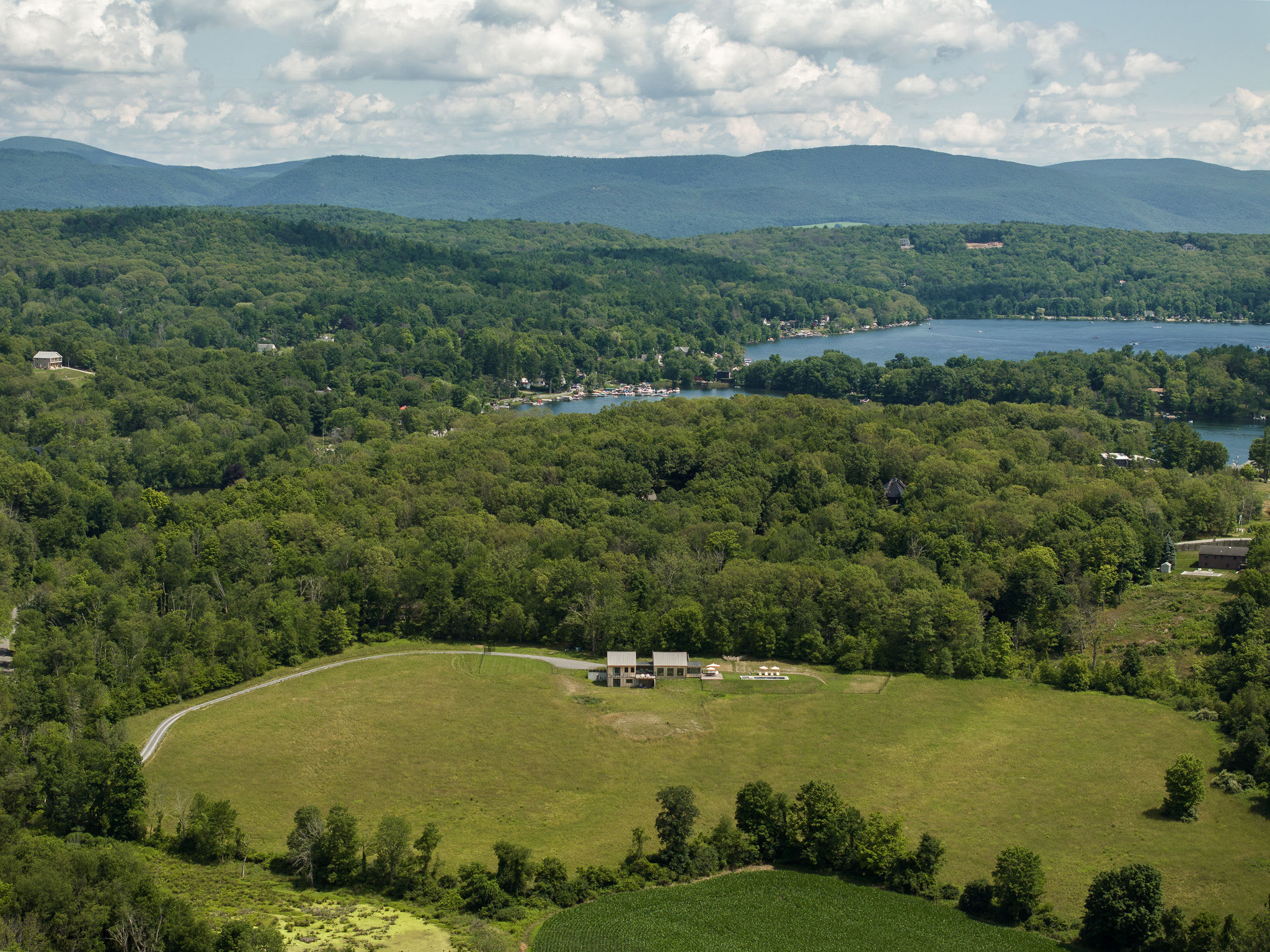 An aerial view showcases a modern house nestled in a clearing amidst lush green forests and rolling hills, with a distant lake and mountains under a partly cloudy sky. A winding driveway leads to the property, which is surrounded by open fields and dense trees.