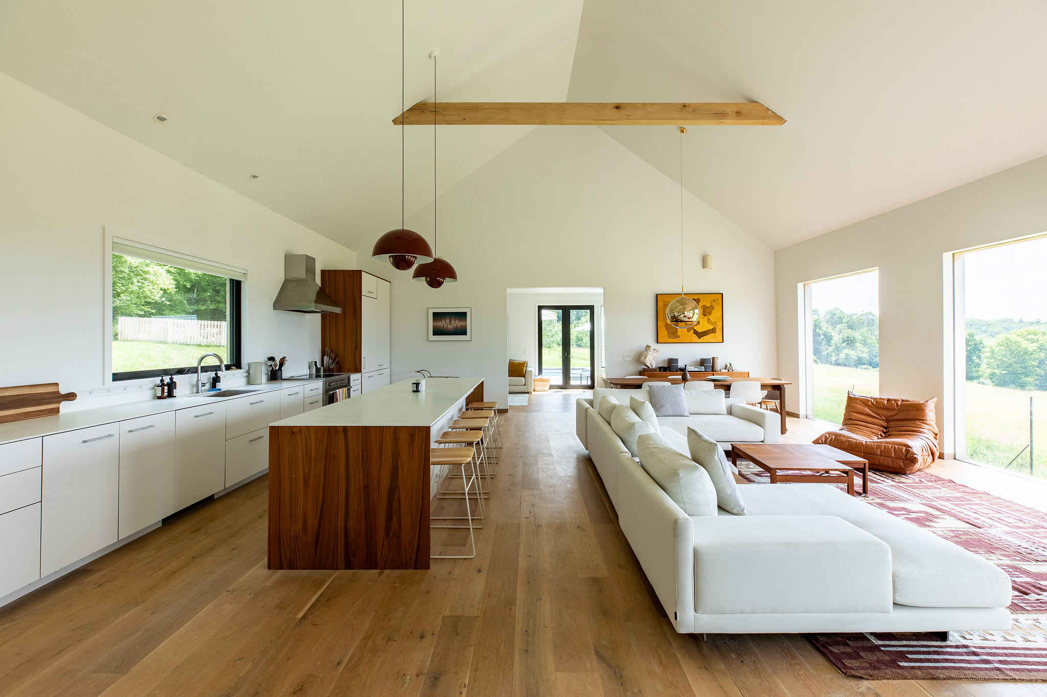 An expansive open-concept living area with vaulted ceilings and light hardwood floors connects the living room, kitchen, and dining area, featuring a large white sectional sofa and panoramic views of the surrounding landscape.