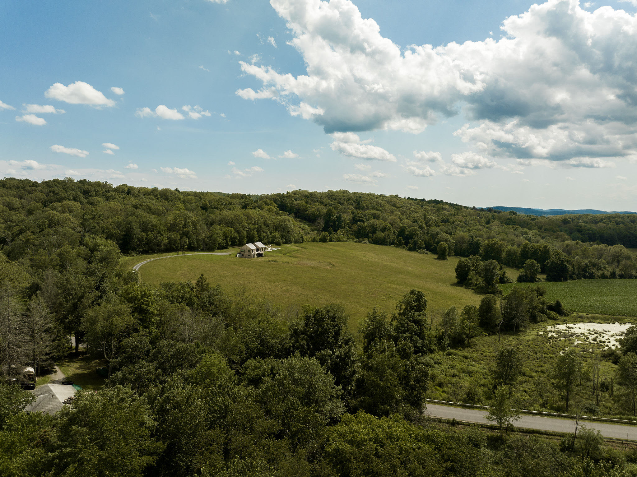 An expansive aerial view showcases a modern house situated in a large, verdant clearing amidst dense green forests and open fields, under a bright blue sky with scattered white clouds.