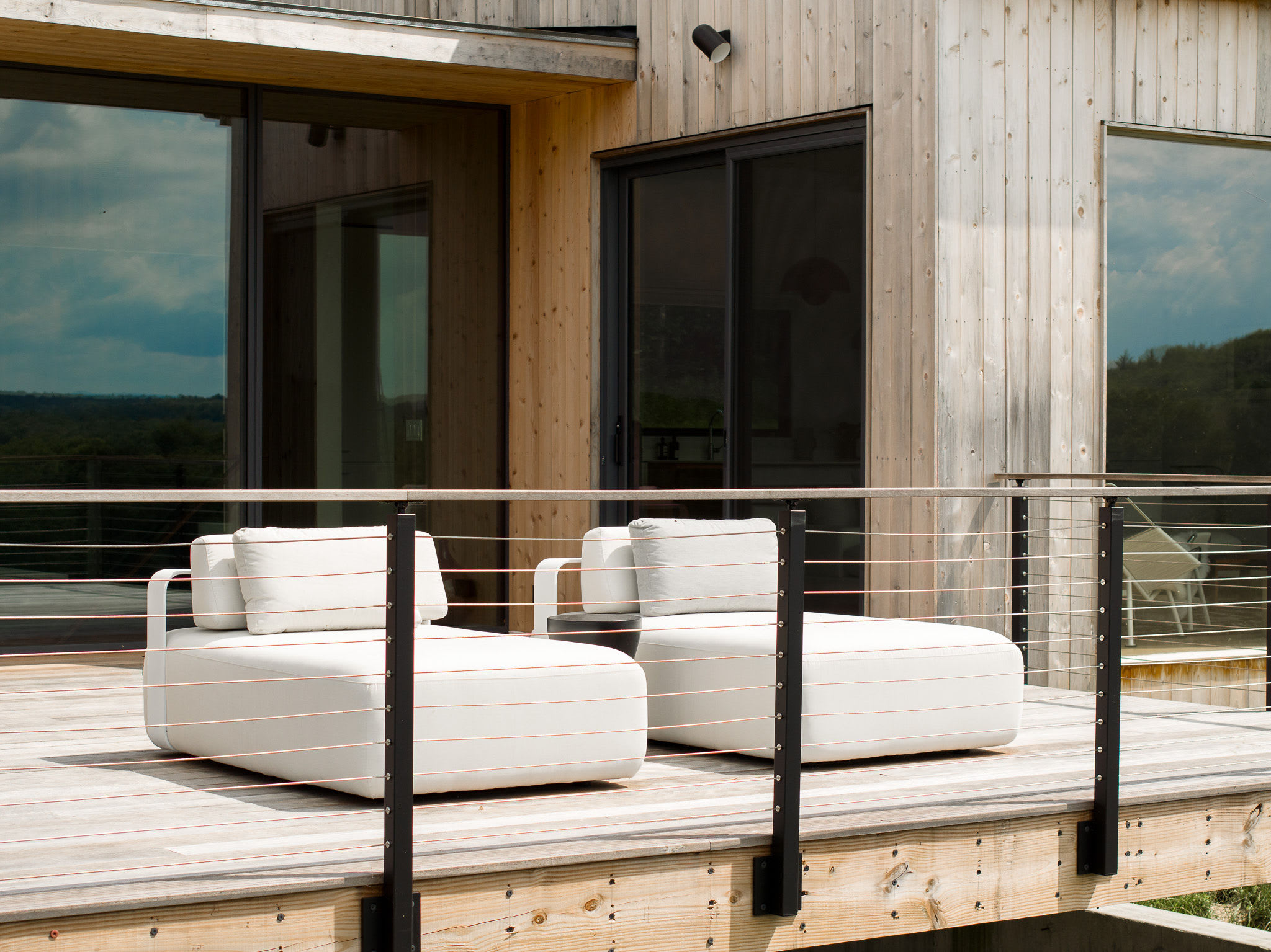 Two contemporary white outdoor lounge chairs are positioned on a light wooden deck with a cable railing, adjacent to a modern house featuring light wooden siding and large black-framed sliding glass doors.