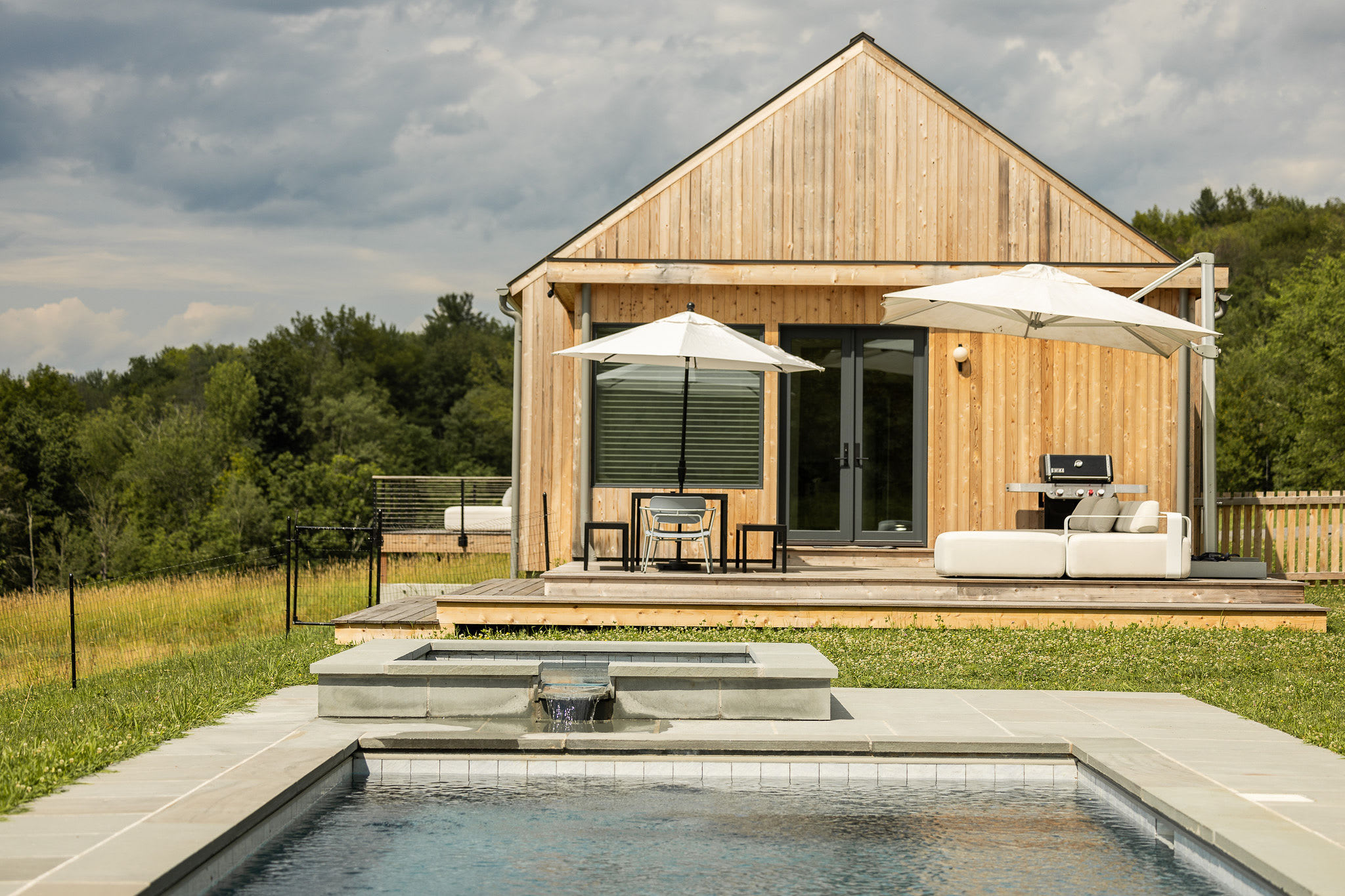The back exterior of a modern wooden house with a gabled roof, featuring a spacious wooden deck with outdoor seating and a grill. A rectangular swimming pool and hot tub are visible in the foreground, surrounded by green grass and distant trees.