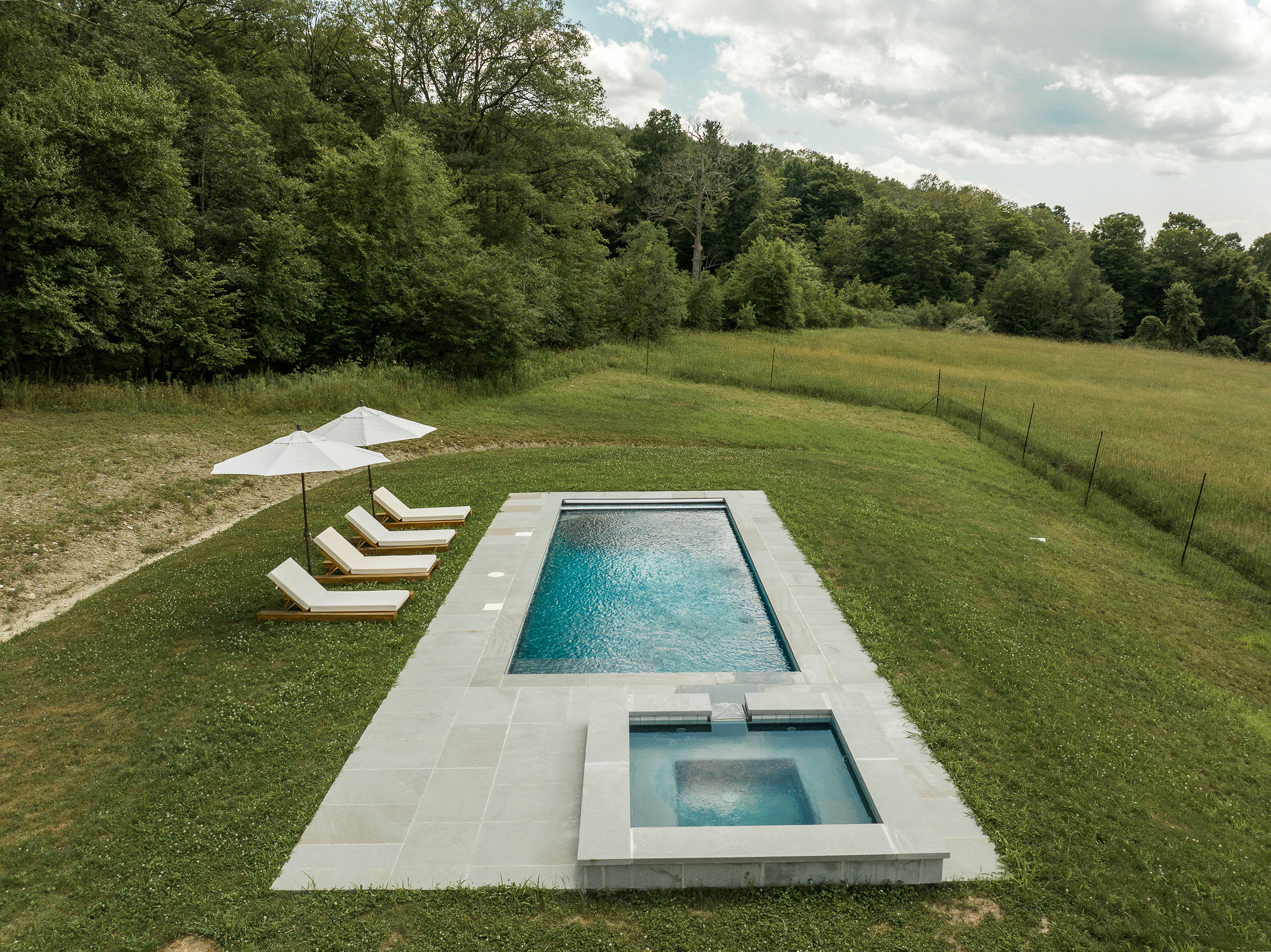 An aerial view shows a rectangular inground swimming pool and a separate square hot tub, both surrounded by grey stone paving, set on a grassy hillside with two white umbrellas and four lounge chairs, backed by a dense forest.