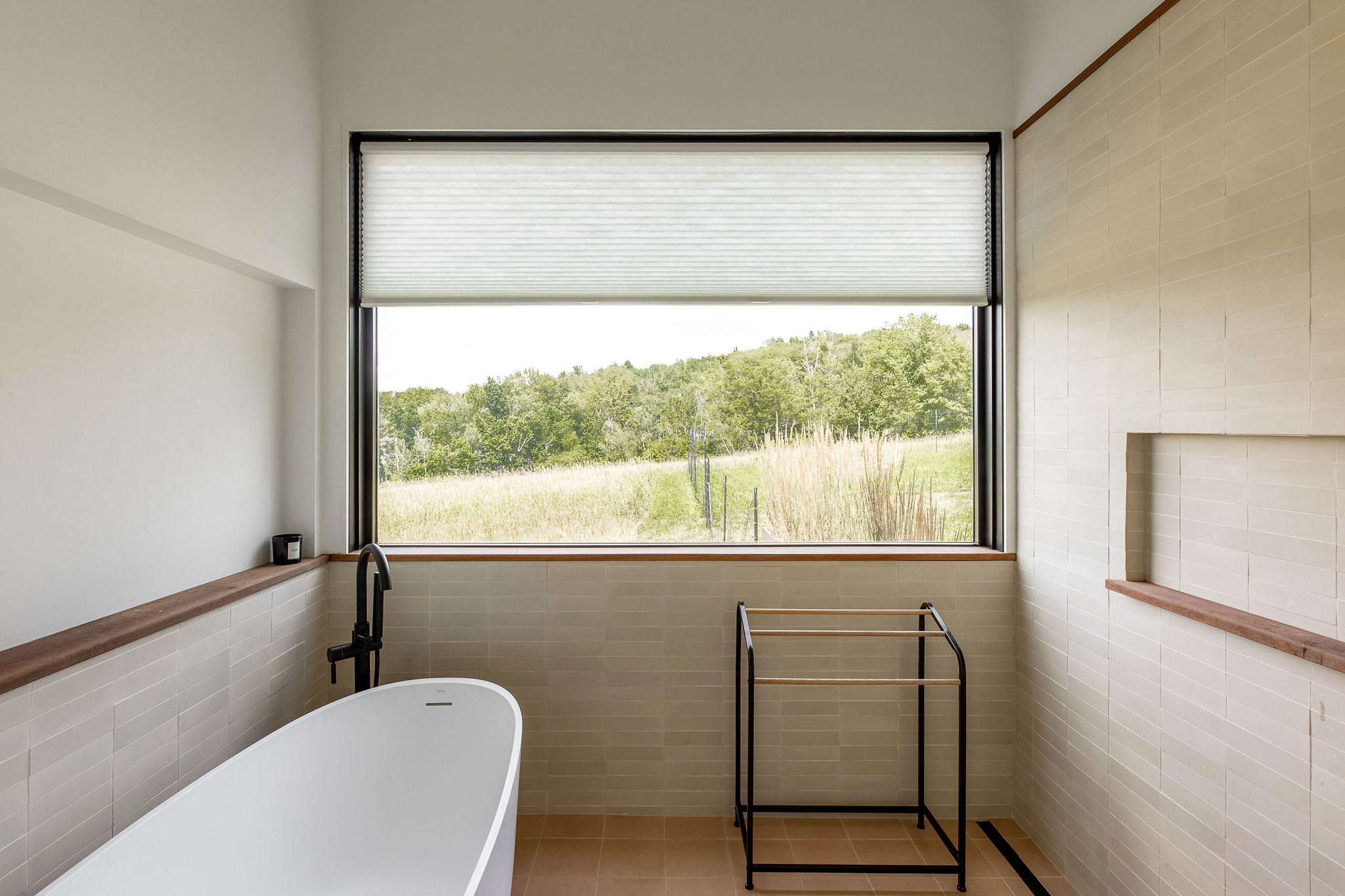 Bright bathroom with a freestanding white bathtub and light-colored subway tiles, featuring a large horizontal window that offers a scenic view of a lush green landscape.