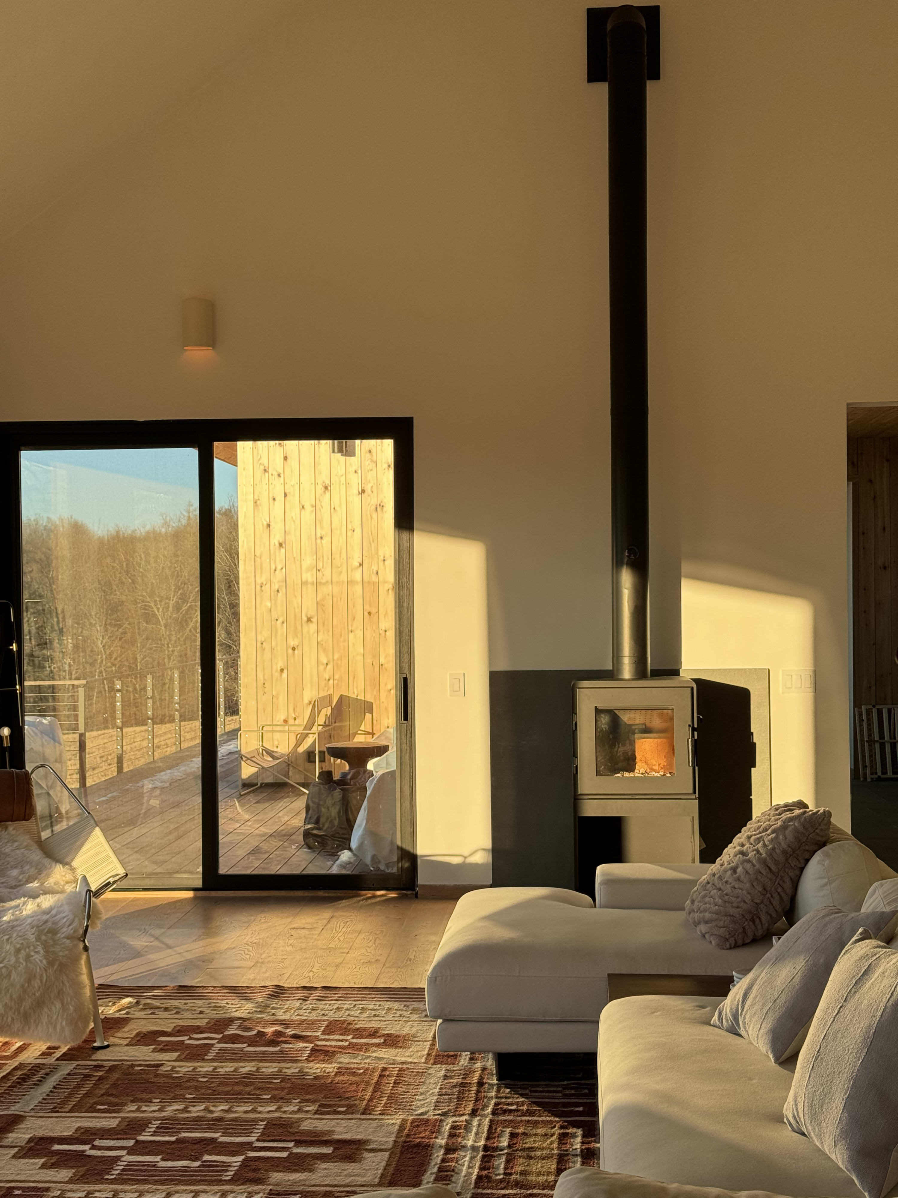 A cozy living room bathed in warm golden hour light features a modern wood-burning stove with a tall black flue, next to a large sliding glass door leading to an outdoor deck. A light-colored sectional sofa with throw pillows is visible, along with a patterned rug.