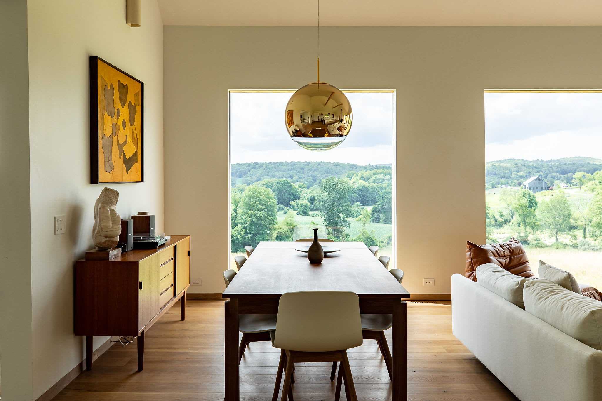 A modern dining area features a long wooden table with six chairs, illuminated by a large gold spherical pendant light, with expansive windows providing scenic views of a green valley and distant hills.