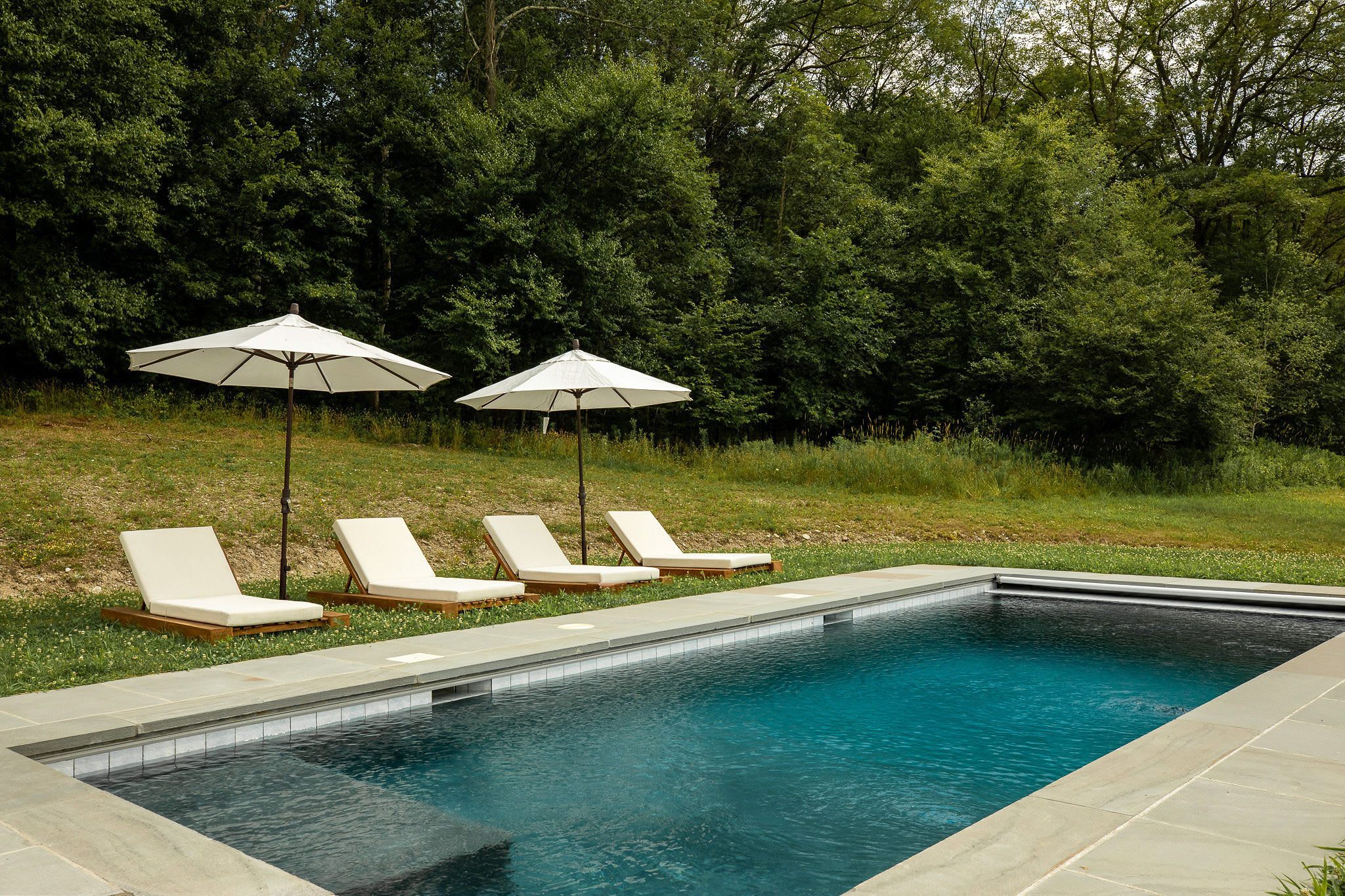 A rectangular inground swimming pool with dark blue water is surrounded by light grey stone coping, with four white lounge chairs and two white umbrellas on a grassy area beside it, backed by lush green trees.