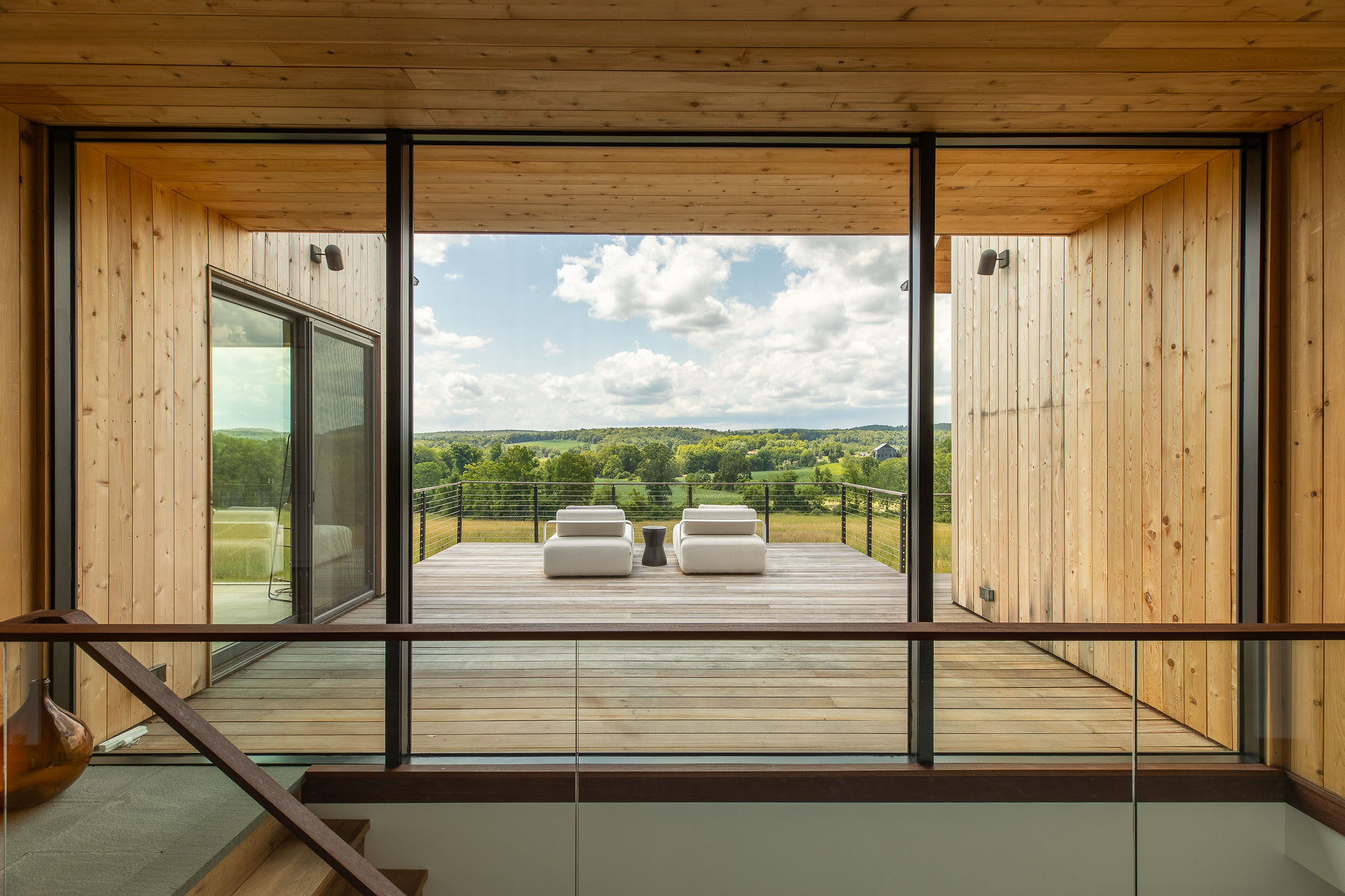 A modern interior balcony overlooks a stunning natural landscape through expansive floor-to-ceiling glass windows and doors, leading to an outdoor deck with two white lounge chairs. The space features light wood paneling on walls and ceiling, and a glass railing.