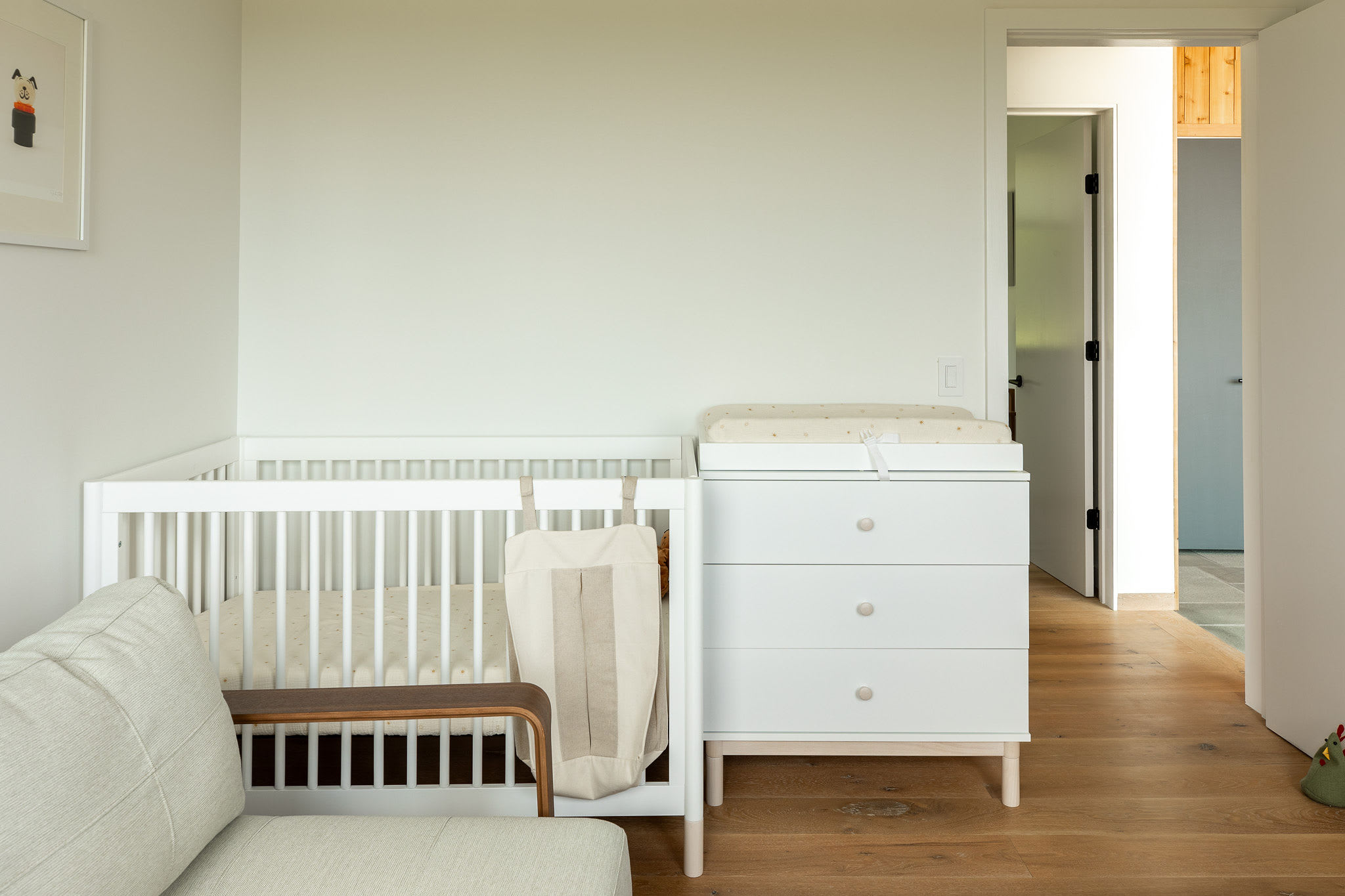 Child-friendly room with a white crib and a matching changing table dresser, positioned next to a light-colored sofa, with hardwood floors and a doorway leading to another part of the house.