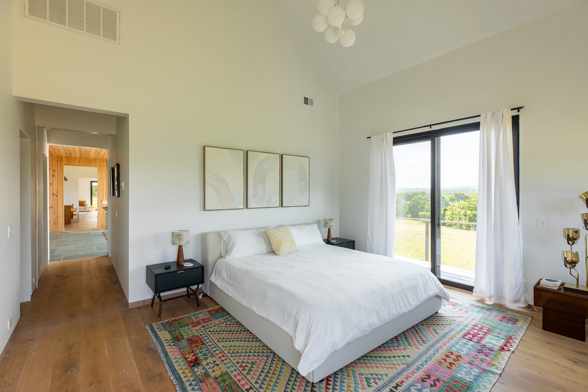 Spacious bedroom with a white platform bed, two dark nightstands, and a vibrant patterned rug, featuring a large glass door leading to outdoor views and modern abstract artwork above the bed.