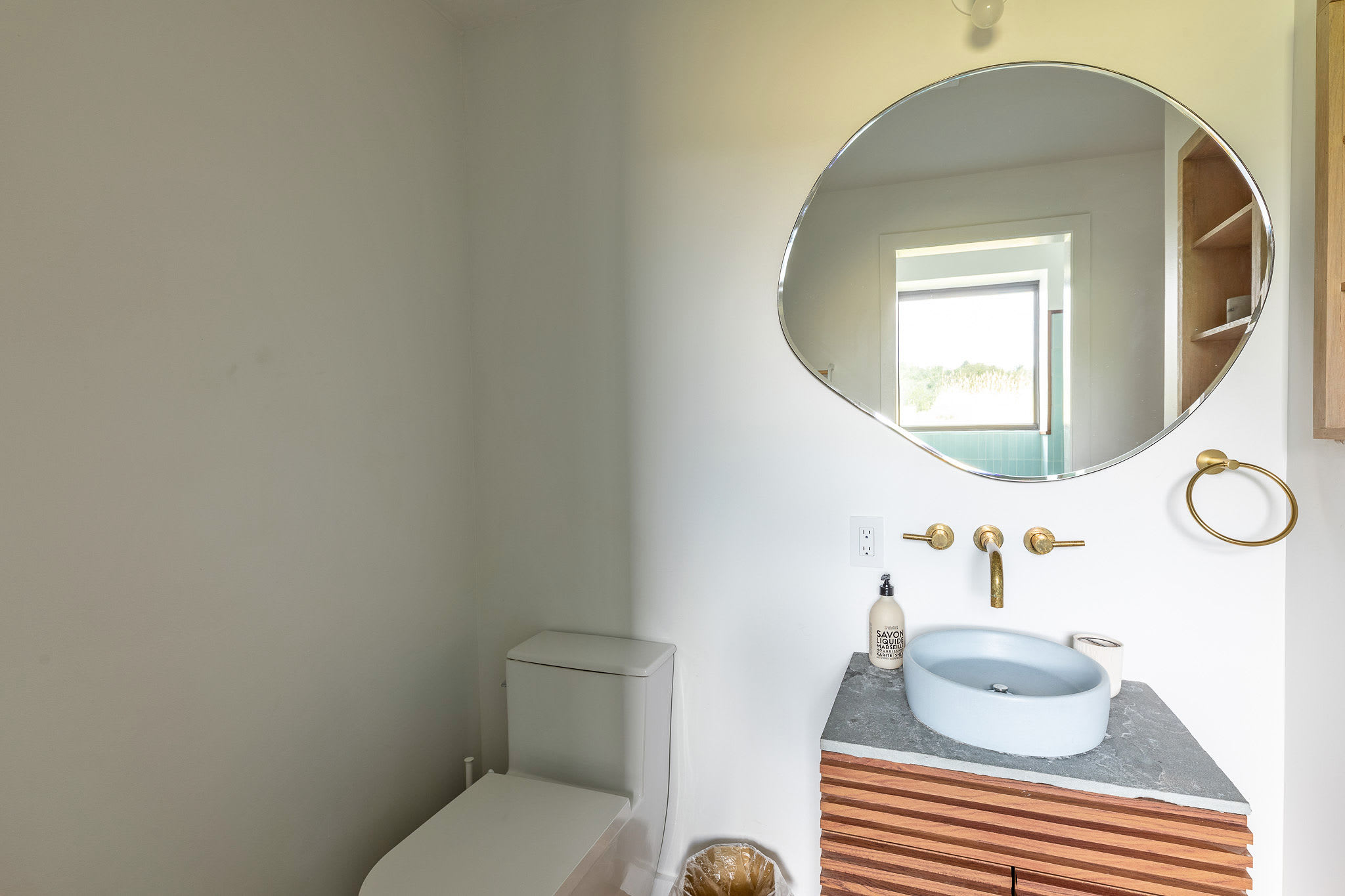 Modern bathroom featuring a white toilet and a unique light blue circular basin sink set on a grey slatted vanity. A large organic-shaped mirror, gold fixtures, and white walls complete the minimalist design.