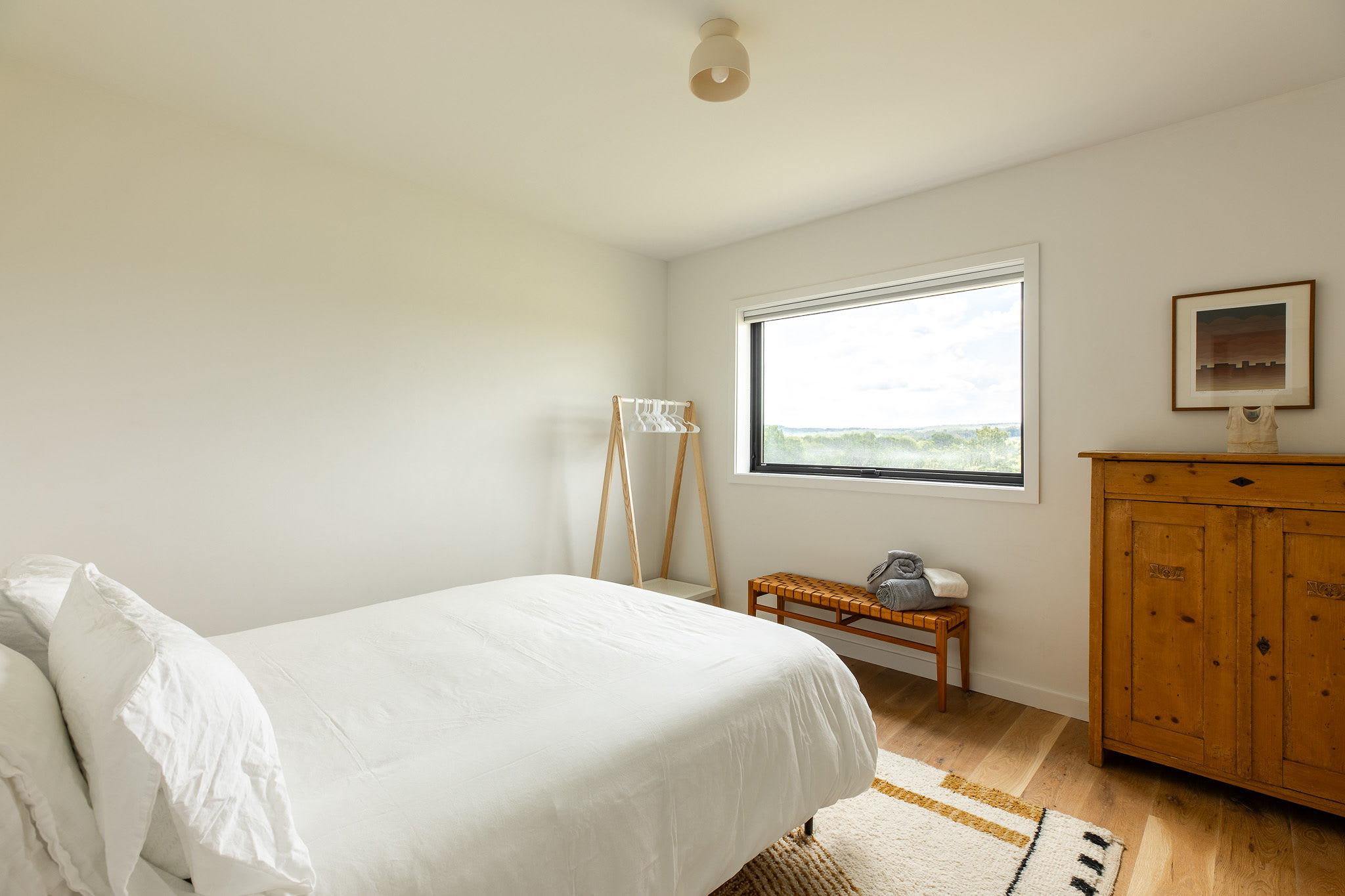 Bright and minimalist bedroom with a queen bed featuring crisp white bedding, light wood flooring, and a large horizontal window overlooking a scenic landscape. A rustic wooden wardrobe and a small bench with a clothes rack add functional elements.
