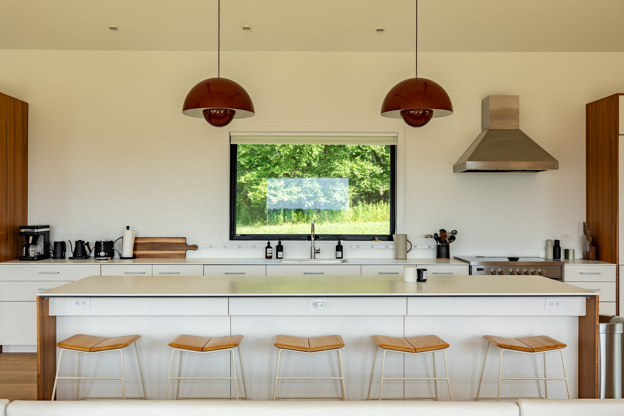 A close-up of the modern kitchen island with a wooden base and white countertop, featuring five light-colored bar stools, a stainless steel range hood, and a large window offering a view of lush green trees.