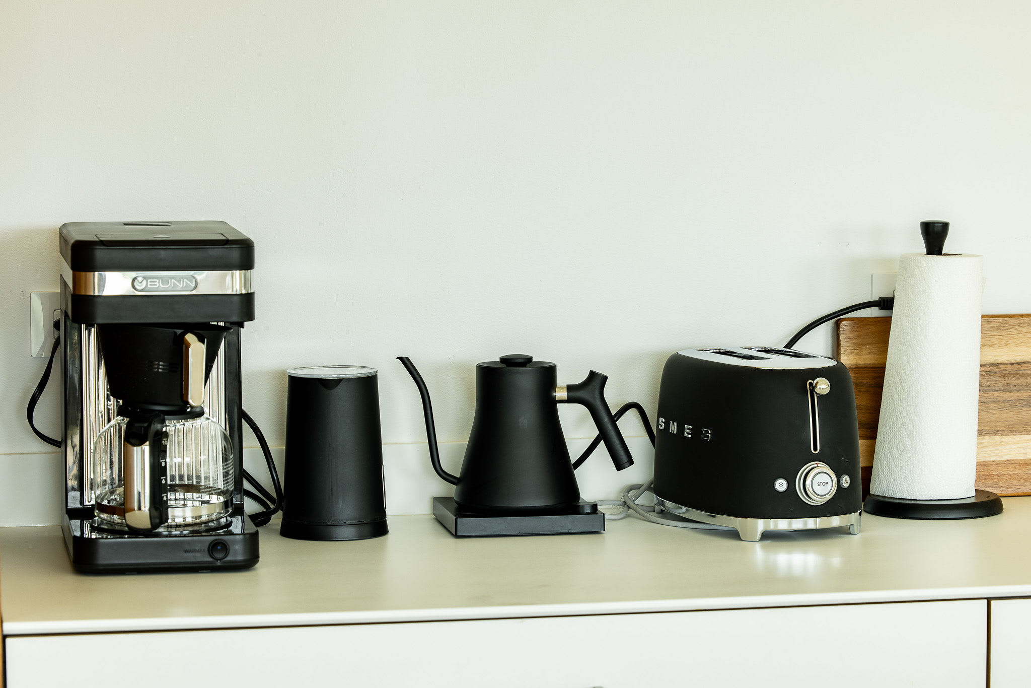 A close-up of a kitchen counter displays a collection of modern black kitchen appliances, including a Bunn coffee maker, a Fellow Stagg EKG kettle, and a Smeg toaster. A roll of paper towels and a wooden cutting board are also visible.
