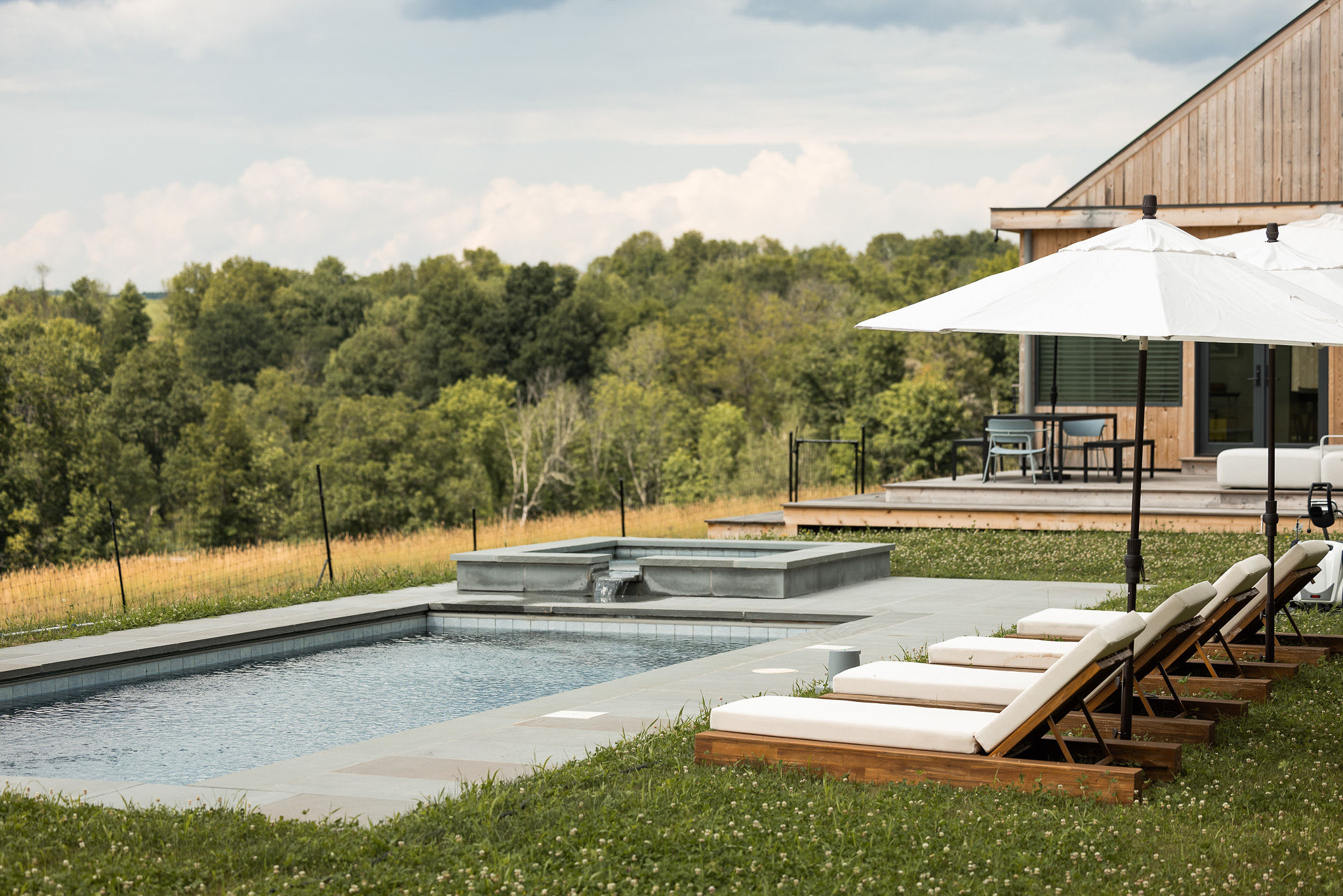 A serene outdoor scene featuring a rectangular swimming pool with grey stone coping, surrounded by a manicured lawn with several wooden lounge chairs. The modern wooden house and a vast green landscape with trees are visible in the background.
