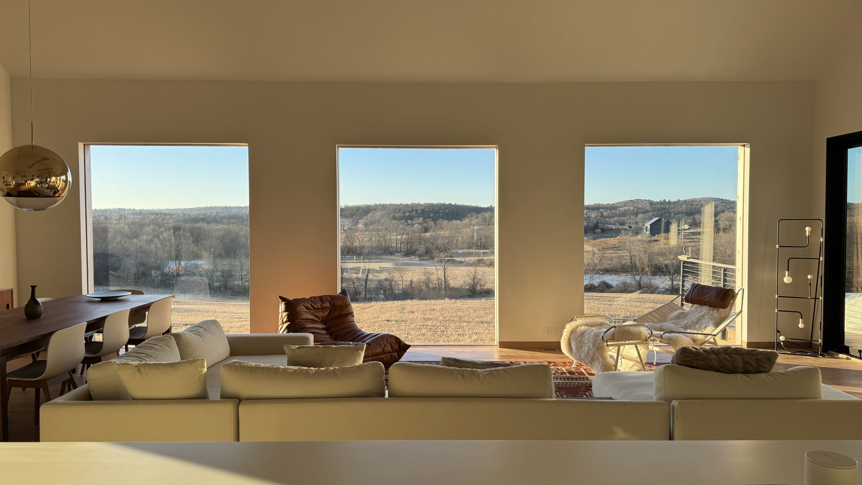 A spacious living room with large rectangular windows offers panoramic views of a snowy, rural landscape under a clear blue sky. The room is furnished with a large white sectional sofa, a brown leather lounge chair, and a dining table in the background.